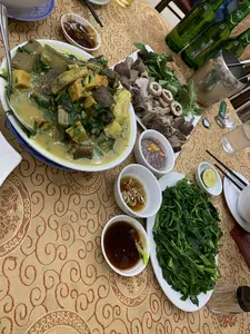 Bối cảnh là một bữa ăn thịnh soạn tại một nhà hàng hoặc một buổi tụ tập gia đình. Trên bàn ăn được trải khăn màu vàng có hoa văn, có nhiều món ăn Việt Nam đặc trưng. Nổi bật là một bát lớn món om có nước dùng màu vàng, bên trong có chuối xanh, đậu phụ, các loại rau thơm và có thể là thịt hoặc lươn, được rắc hạt tiêu bên trên. Bên cạnh đó là một đĩa lòng lợn luộc gồm dồi, gan và các bộ phận khác, ăn kèm với rau thơm. Ngoài ra còn có một đĩa rau xanh luộc. Bữa ăn được phục vụ với nhiều loại nước chấm khác nhau đặt trong các bát nhỏ, bao gồm mắm tôm pha chanh ớt và nước mắm ớt. Đồ uống là bia, với các chai bia màu xanh lá cây có nhãn hiệu "Trúc Bạch" và một xô đá bằng kim loại để giữ lạnh. Bát, đũa và ly thủy tinh được sắp xếp sẵn cho thực khách.。（日本語訳）。背景は、レストランでの豪華な食事、または家族の集まりです。模様のある黄色いテーブルクロスが敷かれた食卓の上には、多くの特徴的なベトナム料理があります。目立っているのは、黄色いスープの煮込み料理が入った大きな鉢で、中には青いバナナ、豆腐、各種香草、そしておそらく肉かウナギが入っており、上には胡椒が振りかけられています。その隣には、茹でた豚の内臓の皿があり、ドイ（腸詰）、レバー、その他の部位が含まれ、香草が添えられています。その他に、茹でた青菜の皿もあります。食事は、小鉢に入れられた様々な種類のつけダレと共に提供され、それにはライムと唐辛子を混ぜたマムトム（エビの発酵調味料）や、唐辛子入りのヌクマム（魚醤）が含まれます。飲み物はビールで、「Trúc Bạch」というブランドの緑色のビール瓶と、冷やすための金属製のアイスペールがあります。碗、箸、ガラスのコップが食事客のために用意されています。