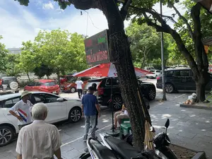 Bản ghi văn bản từ biển hiệu điện tử: "62 Hàng Sắt THĂM HÙNG". Bối cảnh là một vỉa hè trên một con phố vào ban ngày, có nhiều cây xanh lớn. Nhiều người đang đi lại và sinh hoạt trên vỉa hè. Một người đàn ông lớn tuổi mặc áo sơ mi kẻ sọc và một người đàn ông mặc áo phông xanh đang đi bộ. Một người đàn ông khác đội nón cối đang đứng cạnh một chiếc ô tô. Dưới một chiếc ô lớn màu đỏ và trắng có logo "HABECO", một người đang ngồi trên ghế nhựa. Xung quanh có rất nhiều ô tô và xe máy đang đỗ, trong đó có một chiếc xe máy hiệu "WINNER". Một cây chổi được dựa vào gốc cây.。（日本語訳）。電子看板からのテキスト記録：「62 Hàng Sắt THĂM HÙNG」。背景は日中の路上にある歩道で、大きな緑の木がたくさんあります。多くの人々が歩道を行き来し、生活しています。ストライプのシャツを着た年配の男性と、青いTシャツを着た男性が歩いています。ピスヘルメットをかぶった別の男性が、車の横に立っています。「HABECO」のロゴが入った赤と白の大きな傘の下で、一人の人がプラスチックの椅子に座っています。周りには多くの車やバイクが駐車しており、その中には「WINNER」ブランドのバイクが一台あります。一本のほうきが木の根元に立てかけられています。