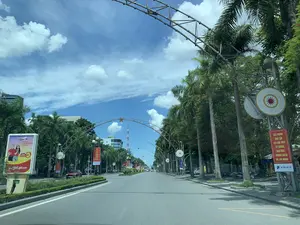 Bức ảnh chụp một con đường lớn, có thể là một đại lộ ở thành phố Thanh Hóa, dưới bầu trời xanh và có nhiều mây trắng. Hai bên đường được trồng nhiều cây cọ cao. Con đường được trang trí bằng các cổng chào bằng kim loại có gắn hình ngôi sao ở trên đỉnh. Có một vài phương tiện như ô tô và xe máy đang di chuyển và đỗ ven đường.。Có nhiều biển quảng cáo và băng rôn có thể đọc được:。- Một biển quảng cáo lớn của hãng hàng không Vietjet Air ở bên trái có nội dung: 'Từ Thanh Hóa → TP. HỒ CHÍ MINH → ĐÀ NẴNG', '12h Vietjet thôi', và 'VietjetAir.com'.。- Các băng rôn màu đỏ treo trên cột. Một băng rôn ghi: 'ĐẨY MẠNH HỌC TẬP VÀ LÀM THEO TƯ TƯỞNG, ĐẠO ĐỨC, PHONG CÁCH HỒ CHÍ MINH。' với logo VNPT bên dưới.。- Một băng rôn khác ghi: 'THI ĐUA LẬP THÀNH TÍCH XUẤT SẮC CHÀO MỪNG KỶ NIỆM 220 NĂM ĐÔ THỊ TỈNH LỴ。' với logo Vinaphone bên dưới.。- Các cột đèn còn được trang trí bằng biểu tượng hình tròn trông giống mặt trống đồng.。- Ở phía xa, có thể thấy các tòa nhà cao tầng, bao gồm một tòa nhà có logo 'BIDV' và một tháp viễn thông.。（日本語訳）。写真は、青空と白い雲の下、タインホア市の大通りと思われる大きな道を写しています。道の両側には、背の高いヤシの木がたくさん植えられています。道は、頂上に星の形が取り付けられた金属製の歓迎ゲートで飾られています。車やバイクなどのいくつかの乗り物が走行したり、道端に駐車したりしています。読み取れる多くの広告看板や横断幕があります：。- 左側には、航空会社ベトジェットエアの大きな広告看板があり、「タインホアから → ホーチミン市 → ダナン」、「12時、ベトジェットだけ」、「VietjetAir.com」という内容が書かれています。- 柱に赤い横断幕が掲げられています。ある横断幕には、「ホーチミンの思想、道徳、スタイルを学び、それに従うことを推進しよう。」と書かれており、その下にはVNPTのロゴがあります。- 別の横断幕には、「省都220周年記念を祝して、優れた功績を競い合おう。」と書かれており、その下にはVinaphoneのロゴがあります。- 街灯の柱は、銅鼓の表面に似た円形のシンボルでも飾られています。- 遠くには、「BIDV」のロゴがあるビルや通信塔などの高層ビルが見えます。