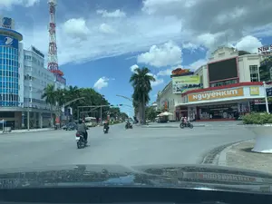 Bức ảnh chụp một ngã tư đường phố sầm uất tại một thành phố ở Việt Nam, có thể là thành phố Thanh Hóa. Nhiều người đang tham gia giao thông, di chuyển bằng xe máy và ô tô. Khung cảnh cho thấy một ngày bình thường tại một trung tâm đô thị.。Các thông tin văn bản có thể nhận dạng bao gồm:。- Tòa nhà bên trái có các biển hiệu lớn ghi 'VNPT' và 'Vinaphone', cùng với một tháp viễn thông cao màu đỏ và trắng. Một biển hiệu khác ghi 'SHOWROOM' và số điện thoại '18001166'.。- Tòa nhà lớn ở góc phải là siêu thị điện máy 'NGUYENKIM'. Phía trên có nhiều biển quảng cáo, bao gồm 'RACERCITY KHU VUI CHƠI TRẺ EM - BÉ BÉ', 'HAYSPORT', 'Nhà Sách Tiền Phong', và một quảng cáo thời trang 'TRUE FASHION SUMMER'. Một biển quảng cáo khác có nội dung 'TOP 10 THƯƠNG HIỆU HÀNG ĐẦU VIỆT NAM NĂM 2023'.。- Một biển tên đường màu xanh lam ghi 'ĐẠI LỘ LÊ LỢI'.。Các vật thể và hoạt động chính trong ảnh:。- Địa điểm: Một ngã tư lớn trong thành phố với nhiều tòa nhà thương mại.。- Hoạt động: Người dân đang lái xe máy và ô tô tham gia giao thông. Một chiếc xe buýt màu vàng cũng có mặt trên đường.。- Vật thể: Xe máy, ô tô, xe buýt, các tòa nhà cao tầng, tháp viễn thông, biển quảng cáo, đèn giao thông, và cây cọ trồng dọc theo đường phố.。（日本語訳）。写真はベトナムのある都市、おそらくタインホア市の賑やかな交差点を撮影したものです。多くの人々がバイクや車で交通に参加しています。この光景は、都市中心部の日常の一日を示しています。認識可能なテキスト情報は以下の通りです：。別の看板には「SHOWROOM」と電話番号「18001166」が書かれています。- 右隅の大きな建物は家電スーパー「NGUYENKIM」です。その上には「RACERCITY KHU VUI CHƠI TRẺ EM - BÉ BÉ」、「HAYSPORT」、「Nhà Sách Tiền Phong」（ティエンフォン書店）、そしてファッション広告「TRUE FASHION SUMMER」など、多くの広告看板があります。別の広告看板には「2023年ベトナムトップ10ブランド」という内容が書かれています。- 青い道路標識には「ĐẠI LỘ LÊ LỢI」（レロイ大通り）と書かれています。写真の中の主な物体と活動：。- 場所：多くの商業ビルが立ち並ぶ市内の大きな交差点。- 活動：人々がバイクや車を運転して交通に参加している。黄色いバスも道路上にいます。- 物体：バイク、車、バス、高層ビル、通信塔、広告看板、信号機、そして通りに沿って植えられたヤシの木。