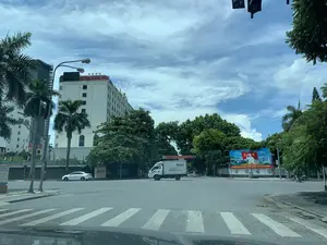 Bối cảnh là một ngã tư đường phố tại thành phố Thanh Hóa vào một ngày trời trong xanh, có mây. Nổi bật trong ảnh là một tòa nhà cao tầng màu trắng với dòng chữ đỏ 'PHƯỢNG HOÀNG HOTEL' và biểu tượng 4 ngôi sao trên nóc. Xung quanh có nhiều cây xanh tươi tốt, bao gồm cả cây cọ. Tại ngã tư có hệ thống đèn giao thông và vạch kẻ đường cho người đi bộ. Về hoạt động, một chiếc xe tải màu trắng đang di chuyển qua ngã tư, trong khi một chiếc ô tô con màu trắng đỗ ở lề đường và một vài chiếc xe máy cũng đang tham gia giao thông. Ngoài ra, có một số biển hiệu và pano: một tấm pano cổ động lớn có hình ảnh Chủ tịch Hồ Chí Minh cùng cờ Đảng và cờ Tổ quốc, với khẩu hiệu 'ĐẨY MẠNH HỌC TẬP VÀ LÀM THEO TƯ TƯỞNG, ĐẠO ĐỨC, PHONG CÁCH HỒ CHÍ MINH。'. Một tấm biển dài màu xanh lá cây có các dòng chữ nhưng chỉ đọc được một phần như '...THÀNH PHỐ THANH HÓA', 'TỔNG PHÁT H...', và '...VĂN HÓA PHẨM - VẬT PHẨM VÀ... THỂ THAO'. Một băng rôn màu đỏ cũng được treo ngang đường nhưng chữ viết khó đọc.。（日本語訳）。背景は、青空に雲が浮かぶ日のタインホア市の街の交差点です。写真の中で目立っているのは、屋上に赤い文字で「PHƯỢNG HOÀNG HOTEL」と書かれ、4つ星のシンボルがある白い高層ビルです。周りにはヤシの木を含む、青々とした多くの緑の木々があります。交差点には信号機と横断歩道があります。交通の様子は、白いトラックが1台交差点を通過しており、一方、白い乗用車が1台路肩に停車し、数台のバイクも通行しています。その他、いくつかの看板やパネルがあります。ホー・チ・ミン主席の肖像、党旗、国旗が描かれた大きなプロパガンダパネルが1枚あり、「ホー・チ・ミンの思想、道徳、スタイルを学び、それに従うことを推進しよう。」というスローガンが書かれています。緑色の長い看板が1枚あり、文字が書かれていますが、「...タインホア市」、「総発H...」、「...文化品 - 物品および...スポーツ」のように一部しか読み取れません。赤い横断幕も道を横切って掲げられていますが、文字は読みにくいです。