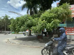 Bối cảnh là một góc đường phố tại Việt Nam vào một ngày nắng, có mây.。**Địa điểm và Cảnh vật:**。Đây là một con đường nhựa tại một khúc cua hoặc bùng binh trong thành phố. Dọc vỉa hè được lát gạch có trồng nhiều cây xanh lớn và cây cọ, tạo bóng mát. Phía sau hàng cây là các cửa hàng ven đường. Trên bầu trời có một cấu trúc trang trí lớn hình vòng cung.。**Hoạt động:**。Hoạt động chính là cảnh giao thông thường ngày. Ở tiền cảnh, một người đang điều khiển một chiếc xe máy tay ga (xe ga) hiệu Honda Air Blade màu đen. Người này mặc một bộ đồ chống nắng màu xanh đậm trùm kín toàn thân và đeo khẩu trang, một trang phục phổ biến để bảo vệ khỏi ánh nắng mặt trời. Ở phía xa, có những người khác cũng đang đi xe máy và ô tô đang lưu thông trên đường.。**Chữ viết và Vật thể:**。*   **Chữ viết:** Có thể đọc được nhiều biển hiệu. Một biển hiệu lớn màu đỏ của tiệm sửa xe máy ghi: "SỬA CHỮA", "XE GA - XE SỐ", tên cửa hàng là "HẢI ĐỘ" tại địa chỉ số "91", và khẩu hiệu "Nơi Gửi Gắn". Các biển hiệu khác có logo "VNPT" và dòng chữ "MÀNH ĐỘNG THÂN THIỆN". Trên chiếc xe máy ở tiền cảnh có chữ "HONDA" và "AIR BLADE".。*   **Vật thể:** Các vật thể nổi bật bao gồm xe máy Honda Air Blade, các loại xe cộ khác, cây xanh, vỉa hè, và các biển hiệu quảng cáo.。（日本語訳）。背景は、晴れて雲のある日のベトナムのある街角です。**場所と風景:**。これは市内のカーブまたはロータリーにあるアスファルトの道です。タイル張りの歩道に沿って、大きな緑の木やヤシの木が多く植えられており、木陰を作っています。並木の向こうには、沿道の店があります。空には大きなアーチ型の装飾的な構造物があります。**活動:**。主な活動は日常の交通風景です。前景では、一人の人が黒いホンダ・エアブレードというスクーターバイク（xe ga）を運転しています。この人は、全身を覆う濃い青色のUVカットウェアを着用し、マスクをしています。これは日光から身を守るための一般的な服装です。遠くには、他にもバイクに乗っている人たちや、道路を通行している自動車がいます。**文字と物体:**。*   **文字:** 多くの看板が読み取れます。あるバイク修理店の大きな赤い看板には、「SỬA CHỮA」、「XE GA - XE SỐ」、店名は「HẢI ĐỘ」、住所は「91」番、そしてスローガン「Nơi Gửi Gắn」と書かれています。他の看板には「VNPT」のロゴと「MÀNH ĐỘNG THÂN THIỆN」という文字があります。前景のバイクには「HONDA」と「AIR BLADE」の文字があります。*   **物体:** 目立つ物体には、ホンダ・エアブレードのバイク、その他の車両、緑の木々、歩道、そして広告看板が含まれます。