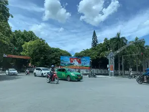 Khung cảnh tại một ngã tư đường phố ở Thanh Hóa vào một ngày nắng, trời xanh có mây. Nhiều phương tiện giao thông bao gồm ô tô và xe máy đang di chuyển trên đường. Mọi người đang tham gia giao thông, một số đang dừng chờ đèn tín hiệu. Hai bên đường có nhiều cây xanh tươi tốt. Ở trung tâm có một tấm biển tuyên truyền lớn và một băng rôn màu đỏ treo ngang đường.。Nội dung chữ viết được ghi lại:。1. Trên tấm biển lớn màu xanh có hình ảnh Chủ tịch Hồ Chí Minh, cờ Đảng, cờ Tổ quốc, cầu Hàm Rồng và một thành phố hiện đại. Bên dưới có dòng chữ: 'ĐẨY MẠNH HỌC TẬP VÀ LÀM THEO TƯ TƯỞNG, ĐẠO ĐỨC, PHONG CÁCH HỒ CHÍ MINH。'.。2. Trên băng rôn màu đỏ treo ngang đường có dòng chữ: 'PHÁT HUY SỨC MẠNH KHỐI ĐẠI ĐOÀN KẾT TOÀN DÂN TỘC, ĐẨY MẠNH TOÀN DIỆN CÔNG CUỘC ĐỔI MỚI VÌ DÂN GIÀU, NƯỚC MẠNH, DÂN CHỦ, CÔNG BẰNG, VĂN MINH。'.。3. Trên cột đỡ kết cấu thép có một biển hiệu nhỏ màu đỏ với logo 'VNPT'.。4. Đèn tín hiệu giao thông đang hoạt động, một bên hiển thị số '20' màu xanh, bên kia hiển thị số '26' màu đỏ.。（日本語訳）。晴れた日、青空に雲が浮かぶタインホア省のある交差点の光景。自動車やバイクを含む多くの交通車両が道路を走行している。人々は交通に参加しており、一部は信号待ちで停止している。道路の両側には青々とした緑の木々が多くある。中央には大きなプロパガンダ看板と、道路を横切って掲げられた赤い横断幕がある。記録された文字の内容：。1. 大きな青い看板には、ホー・チ・ミン主席、党旗、国旗、ハムロン橋、そして近代的な都市のイメージが描かれている。その下には「ホー・チ・ミンの思想、道徳、風格に学び、それらに従うことを推進しよう。」という文言がある。2. 道路を横切って掲げられた赤い横断幕には、「全民族大団結の力を発揮し、豊かな人民、強い国、民主、公正、文明のためにドイモイ（刷新）事業を全面的に推進しよう。」という文言がある。3. 鉄骨構造の支柱には、「VNPT」のロゴが入った小さな赤い看板がある。4. 交通信号が作動しており、一方は緑色で「20」の数字を、もう一方は赤色で「26」の数字を表示している。