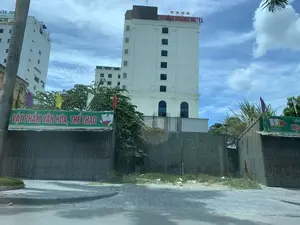 Các dòng chữ ghi lại được từ hình ảnh: Trên nóc tòa nhà cao tầng màu trắng có dòng chữ đỏ "PHƯỢNG HOÀNG HOTEL" và biểu tượng bốn ngôi sao. Một phần của tòa nhà bên cạnh có chữ "NH H". Ở phía trước, có một tấm bạt màu xanh lá cây ghi "VẬT PHẨM VĂN HÓA, THỂ THAO". Một tấm bạt khác bên phải có chữ "SAVINA" và các chữ khác không rõ.。Bối cảnh là một con đường trong đô thị. Nổi bật là tòa nhà khách sạn cao tầng "Phượng Hoàng Hotel" màu trắng. Ở tiền cảnh, có một con đường nhựa và vỉa hè lát gạch. Dọc vỉa hè là các ki-ốt hoặc cửa hàng nhỏ, có vẻ đã đóng cửa với cửa cuốn bằng kim loại. Các cửa hàng này có treo bạt quảng cáo. Giữa các ki-ốt là một khu đất trống được rào lại bằng tường bê tông, có cỏ mọc. Cây xanh và một vài lá cờ nhiều màu sắc cũng xuất hiện trong khung cảnh. Bầu trời xanh và có mây. Không có người hoặc hoạt động cụ thể nào được ghi nhận.。（日本語訳）。画像から記録された文字：白い高層ビルの屋上には、赤い文字で「PHƯỢNG HOÀNG HOTEL」と4つ星のシンボルがあります。隣の建物の一部には「NH H」という文字があります。手前には、「VẬT PHẨM VĂN HÓA, THỂ THAO」（文化・スポーツ用品）と書かれた緑色の防水シートがあります。右側の別の防水シートには「SAVINA」という文字と、その他の不明瞭な文字があります。背景は都市の通りです。目立っているのは、白い高層ホテルの建物「Phượng Hoàng Hotel」です。前景には、アスファルトの道路とタイル張りの歩道があります。歩道沿いにはキオスクや小さな店があり、金属製のシャッターで閉められているようです。これらの店には広告の防水シートが掛けられています。キオスクの間には、コンクリートの壁で囲まれ、草が生えている空き地があります。緑の木々やいくつかのカラフルな旗も風景の中に現れています。空は青く、雲があります。人や特定の活動は記録されていません。