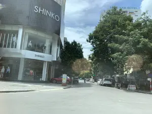Bức ảnh chụp một góc đường phố trong thành phố. Bên trái là một tòa nhà thương mại hiện đại, là cửa hàng thời trang có tên "SHINKO". Tên thương hiệu "SHINKO" được ghi bằng chữ lớn trên mặt tiền và phía trên lối vào. Qua lớp cửa kính, có thể thấy các ma-nơ-canh đang trưng bày quần áo. Bên trong cửa hàng có một tấm biển quảng cáo màu đỏ ghi "QUÀ TẶNG 1 TRIỆU". Dọc theo con đường bên phải, có nhiều cây xanh, ô tô đang di chuyển và đỗ ven đường. Ở phía xa, có một tòa nhà cao tầng màu trắng với dòng chữ "LONG ANH HOTEL". Trên vỉa hè bên phải, có một quầy dịch vụ nhỏ với tấm biển chữ A ghi "CẮT CHÌA KHÓA", "SỬA KHÓA TẠI NHÀ" và số điện thoại "ĐT: 0916.426.785". Một vài người đang đứng gần quầy dịch vụ này.。（日本語訳）。街の一角を撮影した写真です。左側には近代的な商業ビルがあり、「SHINKO」という名前のファッションストアです。ブランド名「SHINKO」は、ファサードと入口の上に大きな文字で書かれています。ガラスのドア越しに、服を陳列しているマネキンが見えます。店内には「QUÀ TẶNG 1 TRIỆU」と書かれた赤い広告看板があります。右側の道沿いには、多くの緑の木々があり、車が走行したり、道端に駐車したりしています。遠くには、「LONG ANH HOTEL」という文字が入った白い高層ビルがあります。右側の歩道には、「CẮT CHÌA KHÓA」、「SỬA KHÓA TẠI NHÀ」、そして電話番号「ĐT: 0916.426.785」と書かれたA型看板のある小さなサービス屋台があります。数人がこのサービス屋台の近くに立っています。