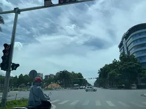 Bối cảnh là một ngã tư đường phố lớn có vòng xuyến tại thành phố Thanh Hóa vào một ngày nắng, trời xanh và có mây. Ở phía trước, một người đi xe máy mặc áo khoác trùm đầu màu xám và đội mũ bảo hiểm màu đỏ đang dừng chờ đèn đỏ. Đèn tín hiệu giao thông đang hiển thị số đếm ngược là '27'. Bên phải là tòa nhà cao tầng hiện đại của ngân hàng BIDV. Giữa vòng xuyến có một tấm biển lớn hình vòm màu đỏ với dòng chữ màu vàng 'ĐẢNG BỘ VÀ NHÂN DÂN THÀNH PHỐ THANH HÓA', dòng chữ bên dưới khó đọc. Nhiều phương tiện khác như ô tô và xe buýt cũng đang lưu thông trên đường. Hai bên đường và trong vòng xuyến được trồng nhiều cây xanh, bao gồm cả cây cọ.。（日本語訳）。背景は、晴れた日のタインホア市にある、ロータリーのある大きな交差点です。空は青く、雲があります。前方では、灰色のフード付きジャケットを着て赤いヘルメットをかぶったバイクの運転手が、赤信号で停止して待っています。交通信号機には「27」というカウントダウンの数字が表示されています。右側にはBIDV銀行の近代的な高層ビルがあります。ロータリーの中央には、黄色い文字で「ĐẢNG BỘ VÀ NHÂN DÂN THÀNH PHỐ THANH HÓA」と書かれた大きな赤いアーチ型の看板があり、その下の文字は読みにくいです。自動車やバスなど、他の多くの車両も道路を通行しています。道路の両側とロータリーの中には、ヤシの木を含む多くの緑の木が植えられています。