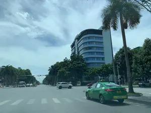 Bối cảnh là một con đường rộng, nhiều làn xe tại một ngã tư trong một thành phố vào một ngày có mây. Hoạt động chính là các phương tiện giao thông đang dừng chờ đèn đỏ. Ở bên phải, một chiếc taxi màu xanh lá cây đang đỗ bên lề đường. Ở giữa ngã tư, một chiếc ô tô SUV màu bạc đang dừng lại. Phía xa là một chiếc xe buýt và nhiều xe máy khác cũng đang chờ tín hiệu giao thông. Các vật thể trong ảnh bao gồm một tòa nhà cao tầng hiện đại với mặt tiền cong và cửa sổ kính màu xanh, cây cọ và các loại cây xanh khác dọc hai bên đường, và hệ thống đèn tín hiệu giao thông. Các cổng chào trang trí hình ngôi sao bắc ngang qua con đường ở phía xa. Thông tin bằng chữ có thể đọc được trên chiếc taxi màu xanh lá cây bao gồm tên hãng 'TAXI MEILINH THANH HÓA', số điện thoại '02373.75.75.75', và số hiệu xe '1055'. Trên cản sau của xe taxi có một decal quảng cáo màu vàng ghi 'ĐÈN LED MPE THIẾT BỊ ĐIỆN'.。（日本語訳）。背景は、ある都市の交差点にある、曇りの日の広くて複数車線のある道路です。主な動きは、交通車両が赤信号で停止して待っていることです。右側には、一台の緑色のタクシーが路肩に停車しています。交差点の中央には、一台の銀色のSUV車が停止しています。遠方には、一台のバスと他の多くのバイクも同じく信号を待っています。写真の中の物体には、湾曲したファサードと青いガラス窓を持つ近代的な高層ビル、道路の両脇に沿ったヤシの木やその他の緑樹、そして信号機システムが含まれます。遠方には、星の形をした装飾的なゲートが道路を横切って架かっています。緑色のタクシーで読み取れる文字情報には、会社名「TAXI MEILINH THANH HÓA」、電話番号「02373.75.75.75」、そして車両番号「1055」が含まれます。タクシーの後部バンパーには、「ĐÈN LED MPE THIẾT BỊ ĐIỆN」と書かれた黄色い広告ステッカーがあります。