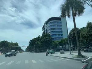 Bức ảnh chụp một con đường lớn, nhiều làn xe tại một thành phố, có thể là thành phố Thanh Hóa, vào một ngày trời có mây. Hoạt động chính là giao thông đô thị. Bên phải đường là một tòa nhà cao tầng hiện đại có kiến trúc cong, tường màu xanh nhạt và cửa sổ kính màu xanh lam; trên đỉnh tòa nhà có chữ "GLOBAL". Trên đường có các phương tiện đang tham gia giao thông, bao gồm một chiếc ô tô màu xám, một chiếc taxi màu xanh lá cây và một số xe máy. Hệ thống đèn tín hiệu giao thông treo phía trên đang bật đèn đỏ. Hai bên đường được trồng nhiều cây xanh, bao gồm cả những cây cọ cao.。（日本語訳）。写真は、曇りの日に、ある都市、おそらくタインホア市の、大きな多車線の道路を写したものです。主な活動は都市交通です。道路の右側には、曲線的な建築様式の近代的な高層ビルがあり、壁は薄い青色で、窓は青いガラスです。ビルの最上部には「GLOBAL」という文字があります。道路上には、灰色の乗用車1台、緑色のタクシー1台、そして数台のバイクを含む車両が通行しています。上に吊り下げられた交通信号機は赤信号が点灯しています。道路の両側には、高いヤシの木を含む多くの緑の木が植えられています。