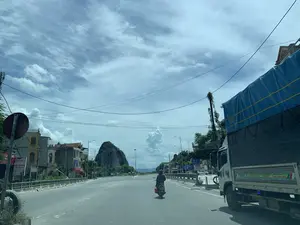 Bức ảnh chụp khung cảnh trên một con đường quốc lộ rộng rãi ở Việt Nam, có thể là tại khu vực Thanh Hóa. Bầu trời trong xanh, có nhiều mây. Con đường khá vắng vẻ, ở giữa có một người đang điều khiển xe máy. Bên phải đường có một chiếc xe tải lớn phủ bạt màu xanh. Hai bên đường là những ngôi nhà dân và cây cối, phía xa nổi bật một ngọn núi đá vôi. Có một cây cầu bê tông ở bên trái.。Các thông tin bằng chữ có thể nhận dạng bao gồm:。- Biển quảng cáo bên trái đường ghi: "BIA VIỆT", "CẦU ĐỒNG", "CƠM BÌNH DÂN". Ngoài ra còn có một biển hiệu nhỏ ghi "PHOTO".。- Biển báo giao thông màu xanh bên phải ghi: "CẦU ĐỒNG", "Km 76 + 450".。- Trên thân xe tải có ghi tên nhà xe "Thành Đạt" và số điện thoại "HOTLINE 0922.604.999".。（日本語訳）。ベトナムの広い国道で撮影された風景写真で、おそらくタインホア省の地域でしょう。空は青く澄んでおり、雲が多いです。道路はかなり閑散としており、中央にバイクを運転している人が一人います。道路の右側には、青いシートで覆われた大きなトラックが一台あります。道路の両側には民家や木々があり、遠くには石灰岩の山が際立っています。左側にはコンクリートの橋があります。識別可能な文字情報は以下の通りです：。- 道路左側の広告看板には、「BIA VIỆT」、「CẦU ĐỒNG」、「CƠM BÌNH DÂN」と書かれています。また、「PHOTO」と書かれた小さな看板もあります。- 右側の青い交通標識には、「CẦU ĐỒNG」、「Km 76 + 450」と書かれています。- トラックの車体には、運送会社名「Thành Đạt」と電話番号「HOTLINE 0922.604.999」が書かれています。
