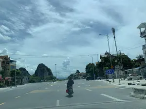 Bức ảnh chụp một khung cảnh giao thông ban ngày trên một con đường lớn, rộng rãi ở khu vực Thanh Hóa. Từ góc nhìn trong một chiếc ô tô, hình ảnh cho thấy một người đang lái xe máy ở giữa đường, mặc áo chống nắng chấm bi trùm kín. Con đường nhựa có nhiều làn xe với vạch kẻ đường và mũi tên chỉ dẫn. Hai bên đường là các ngôi nhà phố, cửa hàng, cây xanh, cột đèn đường và một cột thu phát sóng vi thông. Phía xa là những dãy núi đá vôi đặc trưng dưới bầu trời có nhiều mây. Có một số biển báo và biển hiệu có thể đọc được: Biển chỉ dẫn giao thông màu xanh lam ghi: 'SAO VÀNG-NGHI SƠN 11', 'CAO TỐC BẮC-NAM', 'NÔNG CÔNG 27.8 Km', 'NGÃ BA VOI 7.3 Km' cùng biểu tượng vòng xuyến. Các biển báo phụ khác bao gồm 'CHÚ Ý QUAN SÁT' và biển tam giác 'ĐI CHẬM'. Bên phải đường, có một cửa hàng sửa chữa xe máy tên 'VŨ HƯNG' với biển hiệu 'ENEOS' và 'SỬA CHỮA XE MÁY', cùng một biển hiệu khác ghi 'SƠN LỐP'.。（日本語訳）。写真はタインホア地方の広々とした大きな道路における日中の交通風景を撮影したものです。車内からの視点で、写真には道路の中央でバイクを運転している人が写っており、水玉模様の日焼け防止パーカーで全身を覆っています。アスファルトの道路は多車線で、車線と案内矢印が引かれています。道路の両側には、タウンハウス、店舗、緑の木々、街灯、そして通信用の電波塔があります。遠くには、曇り空の下に特徴的な石灰岩の山々が連なっています。読み取れるいくつかの標識や看板があります。青い交通案内標識には、「SAO VÀNG-NGHI SƠN 11」、「CAO TỐC BẮC-NAM」、「NÔNG CÔNG 27.8 Km」、「NGÃ BA VOI 7.3 Km」と書かれており、ラウンドアバウトのシンボルも描かれています。その他の補助標識には、「CHÚ Ý QUAN SÁT」（注意して観察）と三角形の「ĐI CHẬM」（徐行）標識が含まれます。道路の右側には、「VŨ HƯNG」という名前のバイク修理店があり、「ENEOS」と「SỬA CHỮA XE MÁY」（バイク修理）の看板、そして「SƠN LỐP」（タイヤ塗装）と書かれた別の看板があります。