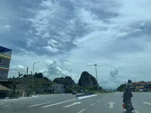 Bức ảnh chụp một con đường rộng lớn, có thể là quốc lộ, tại khu vực Thanh Hóa. Bầu trời trong xanh, có nhiều mây trắng. Phong cảnh nổi bật với những dãy núi đá vôi ở phía sau. Con đường được trải nhựa với các vạch kẻ đường và mũi tên chỉ hướng. Ở giữa đường có dải phân cách. Hai bên đường có các ngôi nhà, cột đèn đường và cột điện. Một người mặc áo chống nắng đang điều khiển xe máy trên đường, phía sau có chở một chiếc xô màu đỏ. Ngoài ra còn có một vài chiếc ô tô đang lưu thông. Có một tấm biển hiệu bên đường ghi chữ 'NEM CHUA THANH HÓA', một đặc sản của vùng này.。（日本語訳）。写真は、タインホア地方の、おそらく国道と思われる広い道を写しています。空は青く澄み渡り、白い雲が多く浮かんでいます。風景は、後方にある石灰岩の山脈が特徴的です。道路はアスファルトで舗装されており、車線や方向を示す矢印が引かれています。道路の中央には中央分離帯があります。道路の両側には、家々、街灯、電柱があります。日よけの上着を着た人がバイクを運転しており、後ろには赤いバケツを積んでいます。その他にも、数台の自動車が走行しています。道端には「NEM CHUA THANH HÓA」と書かれた看板があり、これはこの地域の特産品です。