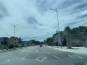 Đây là hình ảnh một con đường nhựa rộng rãi tại một khu vực đô thị hoặc thị trấn ở Thanh Hóa. Con đường có nhiều làn xe, được phân chia bằng các vạch kẻ đường màu trắng, bao gồm mũi tên chỉ hướng và biểu tượng hình thoi. Hai bên đường là những ngôi nhà phố nhiều tầng, một số đang trong quá trình xây dựng. Dọc theo con đường có các cột đèn đường hiện đại và dây điện, dây cáp viễn thông. Có một vài phương tiện đang lưu thông, bao gồm một chiếc ô tô ở phía xa và một người đi xe máy ở bên trái. Vỉa hè được lát gạch và có trồng cây xanh. Bầu trời có mây. Có thể nhìn thấy một số biển hiệu: bên trái có biển hiệu 'Shop Mỹ Phẩm LỢI LINH', và bên phải có biển báo giao thông cho người đi bộ qua đường.。（日本語訳）。これは、タインホア省のある都市部または町にある、広々としたアスファルトの道路の画像です。道路には複数の車線があり、方向を示す矢印やひし形の記号を含む白い車線で区切られています。道路の両側には多層階のタウンハウスが建ち並び、その一部は建設中です。道路沿いには近代的な街灯柱、電線、通信ケーブルがあります。いくつかの車両が通行しており、遠くに一台の自動車、左側にバイクに乗った人が一人います。歩道はタイルで舗装され、植木が植えられています。空は曇っています。いくつかの看板が見えます。左側には「Shop Mỹ Phẩm LỢI LINH」という看板があり、右側には横断歩行者用の交通標識があります。