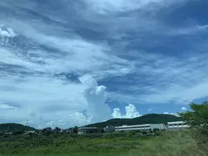 Bức ảnh chụp một phong cảnh ở khu vực nông thôn hoặc ngoại ô vào một ngày có thời tiết đẹp. Bầu trời chiếm phần lớn khung hình, có màu xanh lam với nhiều loại mây: những dải mây ti mỏng ở trên cao và những đám mây tích trắng dày, bồng bềnh, tạo thành các cột lớn vươn lên từ phía chân trời. Phía dưới mặt đất, có một đầm sen rộng với lá xanh và hoa sen trắng. Phía sau đầm sen là một khu dân cư với nhiều ngôi nhà và một tòa nhà lớn, nhiều tầng màu trắng. Xa hơn trong hậu cảnh là những ngọn đồi được bao phủ bởi cây cối xanh tươi. Một cột phát sóng viễn thông cũng có thể được nhìn thấy trên sườn đồi. Không có hoạt động cụ thể nào của con người được ghi lại, và không có văn bản hay biển hiệu nào có thể đọc được.。（日本語訳）。写真は、天気の良い日の田舎または郊外の風景を撮影したものです。空がフレームの大部分を占めており、青色で、様々な種類の雲があります。上空には薄い巻雲の筋があり、地平線からは大きな柱となって立ち上る、厚くふわふわした白い積雲があります。地上には、緑の葉と白い蓮の花が咲く広大な蓮の池があります。蓮の池の後ろには、多くの家と大きな白い多層階の建物がある住宅地があります。さらに奥の背景には、青々とした木々に覆われた丘があります。丘の中腹には通信用の電波塔も見えます。特定の人間の活動は記録されておらず、読み取れる文字や看板もありません。