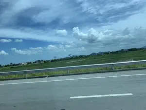 Bức ảnh chụp phong cảnh vùng nông thôn từ một phương tiện đang di chuyển trên đường cao tốc. Phía gần là mặt đường nhựa với vạch kẻ đường và một lan can bảo vệ bằng kim loại. Bên kia lan can là một cánh đồng lúa rộng lớn, xanh mướt, một số khu vực có vẻ như đang ngập nước. Ở phía xa, có thể thấy rải rác một vài ngôi nhà, cây cối và dãy núi trập trùng ở đường chân trời. Có một vài biển quảng cáo lớn được dựng lên ở phía xa nhưng không thể đọc rõ nội dung. Bầu trời trong xanh, có nhiều mây trắng.。（日本語訳）。高速道路を移動中の乗り物から撮影された田園風景の写真です。手前には、路面標示のあるアスファルトの路面と、金属製のガードレールがあります。ガードレールの向こう側には、広大で青々とした水田が広がっており、一部は水に浸かっているように見えます。遠くには、いくつかの家、木々、そして地平線に連なる山々が点在しているのが見えます。空は青く澄んでおり、白い雲が多く浮かんでいます。