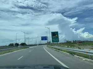 Bức ảnh ghi lại khung cảnh trên một con đường dẫn vào đường cao tốc ở Việt Nam, được chụp từ góc nhìn của người lái xe. Có các biển báo giao thông có thể đọc được:。1. Biển báo màu xanh lam bên trái: "CT.01 MAI SƠN QUỐC LỘ 45 EXPRESSWAY", "LỐI VÀO ENTRANCE".。2. Biển báo màu xanh lá cây bên phải: "NHẬP LÀN MERGE LANE", kèm theo biểu đồ hướng dẫn nhập làn từ bên phải.。3. Biển báo hình tròn viền đỏ: Biển cấm xe thô sơ.。4. Biển báo giới hạn tốc độ: Biển báo hình tròn viền đỏ với số "70" ở giữa, cho biết tốc độ tối đa là 70 km/h.。Cảnh vật trong ảnh là một con đường cao tốc hoặc đường dẫn vào cao tốc, có dải phân cách và hộ lan kim loại. Một vài chiếc ô tô đang lưu thông trên đường. Hai bên đường là khu vực nông thôn với cây cối, đồng ruộng và các cột điện cao thế. Bầu trời trong xanh, có nhiều mây trắng.。（日本語訳）。この写真はベトナムの高速道路への進入路の風景を、運転手の視点から撮影したものです。読み取れる交通標識があります：。1. 左側の青い標識：「CT.01 マイソン－国道45号線高速道路」、「LỐI VÀO ENTRANCE（入口）」。2. 右側の緑色の標識：「NHẬP LÀN MERGE LANE（合流車線）」、右からの合流を示す図が付いています。3. 赤い縁の円形標識：軽車両通行禁止の標識。4. 速度制限標識：中央に「70」と書かれた赤い縁の円形標識で、最高速度が時速70kmであることを示しています。写真の風景は高速道路または高速道路への進入路で、中央分離帯と金属製のガードレールがあります。数台の自動車が道路を走行しています。道路の両側は、木々、田畑、高圧電線の鉄塔がある田園地帯です。空は青く澄んでおり、白い雲が多く浮かんでいます。