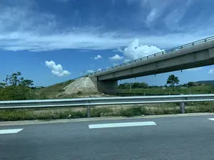 Bức ảnh được chụp từ một phương tiện đang di chuyển, cho thấy khung cảnh một con đường có rào chắn kim loại ở tiền cảnh. Nổi bật trong ảnh là một cây cầu vượt bằng bê tông bắc ngang qua. Bên dưới và xung quanh cây cầu là khu vực đất đai với cây cỏ mọc tự nhiên. Có một biển báo giao thông trên ụ đất gần chân cầu nhưng không thể đọc rõ. Phía xa, qua gầm cầu, có thể nhìn thấy cây cối, cánh đồng và những ngọn đồi. Bầu trời trong xanh và có nhiều mây trắng, cho thấy đây là một ngày nắng đẹp. Toàn bộ khung cảnh mô tả một tuyến đường giao thông hiện đại, có thể là đường cao tốc, đi qua một vùng nông thôn.。（日本語訳）。写真は移動中の乗り物から撮影されたもので、前景に金属製のガードレールがある道路の風景が写っている。写真の中で目立っているのは、横切るように架かるコンクリート製の高架橋である。橋の下と周りには、草木が自然に生えている土地が広がっている。橋の麓近くの土手には交通標識があるが、はっきりと読み取ることはできない。空は青く澄んでおり、白い雲が多く、晴れた美しい日であることを示している。全体の風景は、田舎の地域を通過する現代的な交通路、おそらくは高速道路を描写している。