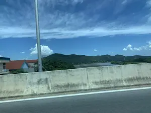 Bức ảnh được chụp từ một phương tiện đang di chuyển, có thể là trên một cây cầu hoặc đường trên cao, nhìn ra khung cảnh bên ngoài. Tiền cảnh là một dải phân cách bằng bê tông và một phần mặt đường nhựa. Ở phía sau dải phân cách là một con sông rộng, hai bên bờ là cây cối xanh tươi. Xa hơn là những dãy núi trập trùng dưới bầu trời xanh và nhiều mây trắng. Có thể thấy một vài ngôi nhà mái ngói đỏ nằm gần bờ sông. Trên mặt sông có những vật thể mờ, có thể là sà lan hoặc thuyền. Trên một cột kim loại ở bên trái có dán nhãn nhưng không thể đọc rõ nội dung.。（日本語訳）。写真は、移動中の乗り物から、おそらく橋または高架道路の上から、外の景色を眺めて撮影されたものです。分離帯の後ろには広い川があり、両岸には青々とした木々が茂っています。その向こうには、青い空と多くの白い雲の下に、起伏のある山々が連なっています。川岸の近くに、赤い瓦屋根の家が数軒見えます。川面には、はしけか船と思われるぼやけた物体があります。左側の金属製の柱にはラベルが貼られていますが、内容ははっきりと読み取れません。
