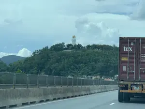 Bức ảnh được chụp từ một phương tiện đang di chuyển trên đường cao tốc. Cảnh vật cho thấy một phần của chiếc xe container màu đỏ ở bên phải, với các dòng chữ như "tex" và "CAUTION 2.9m 9'6" HIGH" trên thân thùng hàng. Con đường có dải phân cách bằng bê tông và hàng rào an toàn. Nhìn về phía xa là một ngọn đồi được bao phủ bởi cây cối xanh tươi. Nổi bật trên đỉnh đồi là một bức tượng đôi lớn màu trắng, có vẻ là một công trình kiến trúc tôn giáo, với phần hào quang màu vàng phía sau. Phía dưới chân đồi có một vài mái nhà dân. Bầu trời trong ảnh có nhiều mây.。（日本語訳）。写真は高速道路を走行中の乗り物から撮影されたものです。風景には、右側に赤いコンテナ車の一部が写っており、コンテナの側面には「tex」や「CAUTION 2.9m 9'6" HIGH」といった文字が見えます。道路にはコンクリート製の中央分離帯とガードレールがあります。遠くには、青々とした木々に覆われた丘が見えます。丘の頂上には、大きな白い二体の像が際立っており、宗教的な建築物のように見え、その後ろには黄色い後光の部分があります。丘の麓には、いくつかの民家の屋根が見えます。写真の空は雲が多いです。