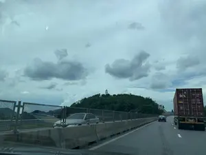 Bức ảnh được chụp từ bên trong một chiếc ô tô đang di chuyển trên đường cao tốc. Khung cảnh cho thấy một đoạn đường cao tốc có nhiều làn xe, với dải phân cách bê tông và hàng rào lưới kim loại. Trên đường có các phương tiện đang lưu thông, bao gồm một chiếc xe đầu kéo chở container ở làn bên phải và một vài chiếc xe ô tô con. Ở phía xa, có một ngọn đồi được bao phủ bởi cây xanh, trên đỉnh đồi có một bức tượng lớn màu trắng. Bầu trời u ám và đầy mây. Trên thùng container màu đỏ có thể đọc được các thông tin sau: logo "tex", mã số "TXGU 557092 0 45G1", cảnh báo "CAUTION 2.9m 9'6" HIGH", và các thông số về trọng lượng như "MAX. WT.", "TARE WT.", "PAYLOAD", "CU. CAP.".。（日本語訳）。写真は高速道路を走行中の車内から撮影されたものです。風景には、複数の車線がある高速道路の一区間が写っており、コンクリートの中央分離帯と金網のフェンスがあります。道路上には車両が走行しており、右車線にはコンテナを運ぶトレーラートラック1台と、数台の乗用車が含まれています。遠くには緑の木々に覆われた丘があり、丘の上には大きな白い像があります。空は曇っており、雲に覆われています。赤いコンテナの上には、以下の情報を読み取ることができます：「tex」のロゴ、識別番号「TXGU 557092 0 45G1」、警告「CAUTION 2.9m 9'6" HIGH」、そして「MAX. WT.」、「TARE WT.」、「PAYLOAD」、「CU. CAP.」といった重量に関する諸元。