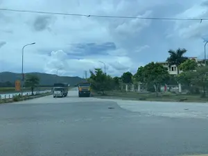 これは、農村部または郊外にある三叉路の光景です。空は曇っています。遠くの道路には、白い箱型トラック1台と黄色いダンプカー1台の、2台のトラックがいます。道路の左側には川または湖があり、その後ろには山脈が広がっています。右側には緑の木々が多く植えられた土地があり、大きな建物が1つあります。屋根に赤地に金色の星のシンボルがあるため、政府機関の建物かもしれません。道路に沿って街灯が設置されています。左側の街灯には赤と黄色の交通標識が取り付けられており、「飲酒したら運転しない」という内容が書かれています