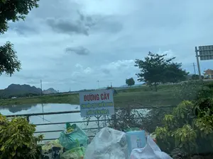 Bức ảnh chụp một khung cảnh ven đường ở vùng nông thôn, có thể được chụp từ bên trong một chiếc ô tô. Ở trung tâm có một tấm biển báo nền trắng, viền xanh với nội dung chữ đỏ "ĐƯỜNG CÂY" và chữ xanh "CHI HỘI CỰU CHIẾN BINH", "THÔN TIẾN HÓA 2". Ngay phía trước và xung quanh tấm biển là một đống rác lớn gồm nhiều túi ni lông và rác thải khác. Cảnh vật phía sau tấm biển bao gồm một con sông hoặc ao, hồ nước. Bên kia bờ là một con đường có cây cối, đồng cỏ và một chiếc ô tô màu trắng đang di chuyển. Ở phía xa là những dãy núi dưới bầu trời nhiều mây, u ám.。（日本語訳）。写真は、田舎の道端の風景を写したもので、車内から撮影された可能性があります。中央には、白い背景に青い縁取りの看板があり、赤い文字で「ĐƯỜNG CÂY」、青い文字で「CHI HỘI CỰU CHIẾN BINH」、「THÔN TIẾN HÓA 2」と書かれています。看板のすぐ手前とその周りには、多くのビニール袋やその他の廃棄物からなる大きなゴミの山があります。看板の後ろの景色には、川または池、湖が含まれています。対岸には、木々や草原のある道があり、白い車が走っています。遠くには、曇り空の下に山々が連なっています。