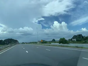 Bức ảnh được chụp từ góc nhìn của người ngồi trong xe ô tô, đang di chuyển trên một con đường cao tốc hoặc quốc lộ rộng rãi có nhiều làn xe. Con đường được trải nhựa phẳng, có vạch kẻ đường màu trắng rõ ràng. Bên phải đường có hàng rào chắn bằng kim loại. Cảnh vật hai bên đường là vùng nông thôn đặc trưng với những dãy núi đá vôi, cây cối xanh tươi và một con sông hoặc hồ nước chạy dọc theo con đường. Bầu trời có nhiều mây, với những đám mây trắng và xám xen kẽ trên nền trời xanh. Bên lề đường có một biển báo giao thông nguy hiểm hình tam giác màu vàng, viền đỏ, cảnh báo đoạn đường bị hẹp ở cả hai bên. Ở phía xa có thể nhìn thấy một vài ngôi nhà và một cột thu phát sóng viễn thông.。（日本語訳）。写真は、複数車線のある広々とした高速道路または国道を走行中の自動車に乗っている人の視点から撮影されたものです。道路は平らに舗装されており、白い車線がはっきりと引かれています。道路の右側には金属製のガードレールがあります。道路の両側の景色は、石灰岩の山々、青々とした木々、そして道路に沿って流れる川または湖がある、特徴的な田園地帯です。空は雲が多く、青空を背景に白と灰色の雲が点在しています。路肩には、黄色い三角形に赤い縁取りの危険を示す道路標識があり、両側から道幅が狭くなることを警告しています。遠くには、数軒の家と通信用の電波塔が見えます。