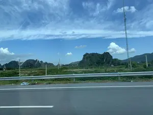 Bức ảnh chụp phong cảnh vùng nông thôn nhìn từ một phương tiện đang di chuyển trên đường. Bầu trời trong xanh với những đám mây trắng. Ở phía xa là những dãy núi đá vôi được cây cối bao phủ. Gần hơn là những cánh đồng xanh, cột điện và một tháp viễn thông cao ở phía bên phải. Ở tiền cảnh là một con đường nhựa có vạch kẻ trắng và một hàng rào chắn bằng kim loại chạy dọc theo lề đường. Không có hoạt động cụ thể nào của con người được nhìn thấy, ngoại trừ việc di chuyển trên đường.。（日本語訳）。道路を移動中の乗り物から見た、田園地帯の風景を撮影した写真です。空は青く澄み渡り、白い雲が浮かんでいます。遠くには、木々に覆われた石灰岩の山々があります。手前には緑の田畑、電柱、そして右側には高い通信塔があります。前景には、白い線が引かれたアスファルトの道路と、路肩に沿って続く金属製のガードレールがあります。道路上の移動を除いて、特定の人間活動は見られません。