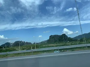 Bức ảnh chụp lại khung cảnh vùng nông thôn ở Thanh Hóa, nhìn từ một phương tiện đang di chuyển trên đường. Ở tiền cảnh là một phần của con đường nhựa với vạch kẻ trắng và lan can hộ lan bằng kim loại. Phía sau lan can là những cánh đồng xanh. Ở hậu cảnh là những dãy núi đá vôi, một số ngọn núi có dấu hiệu bị khai thác đá. Một cột tháp viễn thông cao có thể được nhìn thấy ở phía bên phải. Bầu trời trong xanh, điểm xuyết những đám mây trắng và các vệt trắng do máy bay để lại. Không có hoạt động cụ thể của con người và không có văn bản nào có thể đọc được trong ảnh.。（日本語訳）。この写真は、道路を移動中の乗り物から見た、タインホア省の田舎の風景を撮影したものです。前景には、白い線が引かれたアスファルト道路の一部と、金属製のガードレールがあります。ガードレールの後ろには緑の田畑が広がっています。背景には石灰岩の山々があり、いくつかの山には石を採掘した形跡が見られます。右側には高い通信塔が見えます。空は青く澄んでおり、白い雲と飛行機が残した白い筋が点在しています。特定の人の活動はなく、写真の中には読み取れる文字もありません。