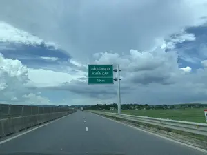 Bức ảnh chụp cảnh một con đường cao tốc, nhìn từ trong xe ô tô. Ở bên trái đường có một biển báo giao thông màu xanh lá cây với dòng chữ trắng: "DẢI DỪNG XE KHẨN CẤP" và "1 Km", báo hiệu rằng còn 1 km nữa sẽ đến dải dừng xe khẩn cấp. Con đường được trải nhựa, có vạch kẻ đường màu trắng. Hai bên đường là dải phân cách bê tông và lan can bảo vệ. Bên phải là một cánh đồng lúa xanh rộng lớn. Phía xa là cảnh làng mạc với nhà cửa, cây cối và những dãy núi. Bầu trời nhiều mây, có cả mây trắng và mây xám.。（日本語訳）。自動車の中から見た高速道路の風景を写した写真です。道路の左側には、白い文字で「DẢI DỪNG XE KHẨN CẤP」（緊急停車帯）と「1 Km」と書かれた緑色の交通標識があり、1km先に緊急停車帯があることを示しています。道路は舗装されており、白い車線が引かれています。道路の両側には、コンクリートの中央分離帯とガードレールがあります。右側には広大な緑の田んぼが広がっています。遠くには、家々、木々、そして山脈のある村の風景が見えます。空は雲が多く、白い雲と灰色の雲の両方があります。