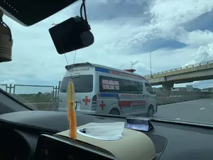 Bức ảnh được chụp từ góc nhìn bên trong một chiếc ô tô đang di chuyển trên đường cao tốc. Phía trước là một chiếc xe cứu thương (ambulance) màu bạc đang chạy cùng chiều. Trên thân xe cứu thương có ghi các dòng chữ: 'TRUNG TÂM VẬN CHUYỂN CẤP CỨU', khẩu hiệu 'NHANH CHÓNG - AN TOÀN - HIỆU QUẢ', chữ 'AMBULANCE', 'STOP', và dòng chữ nhỏ hơn 'Phục vụ 24/24 không có ngày nghỉ'. Ngoài ra còn có hai số điện thoại: '0983.129.408' và '0965.121 115'. Chiếc xe được trang bị đèn hiệu trên nóc và có biểu tượng chữ thập đỏ. Khung cảnh xung quanh là một con đường cao tốc hiện đại với dải phân cách, hàng rào bảo vệ và một cây cầu vượt ở phía xa. Bầu trời nhiều mây. Trong cabin ô tô, có thể thấy một phần táp lô, hộp khăn giấy, và một camera hành trình được gắn trên kính chắn gió.。（日本語訳）。写真は、高速道路を走行中の車内からの視点で撮影されたものです。前方には、同じ方向に走っている銀色の救急車（ambulance）がいます。救急車の車体には、「TRUNG TÂM VẬN CHUYỂN CẤP CỨU」（救急搬送センター）、スローガン「NHANH CHÓNG - AN TOÀN - HIỆU QUẢ」（迅速 - 安全 - 効果的）、「AMBULANCE」、「STOP」、そしてより小さな文字で「Phục vụ 24/24 không có ngày nghỉ」（24時間年中無休で対応）と書かれています。さらに、2つの電話番号「0983.129.408」と「0965.121 115」もあります。この車両は屋根に警告灯を装備し、赤十字のシンボルが付いています。周囲の風景は、中央分離帯、ガードレール、そして遠くに陸橋がある近代的な高速道路です。空は曇っています。車のキャビン内には、ダッシュボードの一部、ティッシュボックス、そしてフロントガラスに取り付けられたドライブレコーダーが見えます。