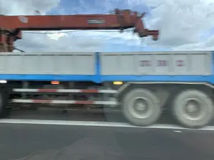 Bức ảnh chụp một chiếc xe tải cẩu đang di chuyển nhanh trên đường nhựa, được chụp từ một phương tiện khác cũng đang di chuyển, gây ra hiệu ứng mờ nhòe do chuyển động. Bối cảnh là một con đường có vạch kẻ trắng, dưới bầu trời xanh có nhiều mây. Chiếc xe tải có cần cẩu màu đỏ, thùng xe màu xám và khung xe màu xanh dương. Dọc bên hông xe có các dải phản quang màu đỏ và trắng. Có thể nhận thấy một dãy số màu sẫm trên thành thùng xe, tuy bị mờ nhưng có vẻ là '078 51 23'. Trên cần cẩu cũng có chữ viết trên một nhãn dán nhưng không thể đọc được.。（日本語訳）。この写真は、アスファルトの道路を高速で移動しているクレーン付きトラックを、同じく移動中の別の車両から撮影したもので、モーションブラー効果が生じています。背景は、白い線が引かれた道路で、上には雲の多い青空が広がっています。トラックは、赤いクレーン、灰色の荷台、そして青いシャーシを備えています。車両の側面には、赤と白の反射テープが貼られています。荷台の側面には、暗い色の数字の列が見て取れますが、ぼやけているものの「078 51 23」のように見えます。