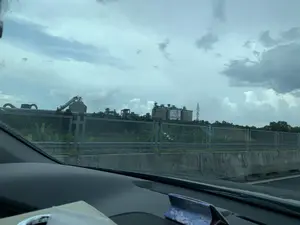 Bức ảnh được chụp từ bên trong một chiếc ô tô đang di chuyển, nhìn ra khung cảnh bên ngoài qua cửa sổ. Ở phía xa là một nhà máy công nghiệp lớn, có thể xác định là nhà máy xi măng qua biển hiệu lớn màu trắng có chữ đỏ 'XI MĂNG DUYÊN HẢI' và logo phía trên, được gắn trên một trong các silo. Nhà máy bao gồm nhiều silo bê tông, hệ thống băng tải và các kết cấu kim loại khác. Ngăn cách giữa con đường và khu vực nhà máy là một bức tường chắn bê tông và một hàng rào lưới thép. Phía sau hàng rào có cây cối, bao gồm cả cây chuối. Bầu trời u ám và đầy mây. Ở phía trước, có thể thấy một phần bảng điều khiển của xe ô tô.。（日本語訳）。写真は移動中の車内から窓越しに外の景色を撮影したものです。遠くには大きな工業工場があり、サイロの一つに取り付けられた、上にロゴがあり赤い文字で「XI MĂNG DUYÊN HẢI」と書かれた大きな白い看板からセメント工場だと特定できます。工場は多くのコンクリートサイロ、コンベアシステム、その他の金属構造物で構成されています。道路と工場エリアの間は、コンクリートの擁壁と金網フェンスで隔てられています。フェンスの後ろには、バナナの木を含む木々があります。空はどんよりとしており、雲に覆われています。手前には、車のダッシュボードの一部が見えます。