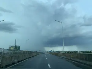 Bối cảnh là một con đường cao tốc hoặc cầu cạn, được chụp từ góc nhìn bên trong một chiếc xe. Mặt đường nhựa bị ướt, cho thấy trời vừa mưa hoặc sắp mưa. Bầu trời u ám, bao phủ bởi những đám mây xám dày đặc. Ở phía xa, có thể thấy rõ một cột mưa lớn đang trút xuống một khu vực, báo hiệu một cơn giông bão.。Hai bên đường là dải phân cách bằng bê tông và hệ thống đèn đường cao áp. Một chiếc ô tô màu trắng đang di chuyển ở phía xa. Bên trái đường có một tấm biển quảng cáo lớn với hình ảnh phong cảnh thiên nhiên. Mặc dù một phần chữ bị mờ, có thể nhận ra các từ như "NINH BÌNH - ĐIỂM ĐẾN..." và tên một công ty "NHỰA HOÀNG THANH NGA". Ở phía xa hơn cũng có các biển báo giao thông màu xanh nhưng không thể đọc được nội dung. Bên phải đường là cảnh quan nhà cửa và cây cối của một khu vực dân cư. Hoạt động chính là việc các phương tiện đang lưu thông trên đường trong điều kiện thời tiết xấu.。（日本語訳）。背景は高速道路または高架橋で、車内からの視点で撮影されています。アスファルトの路面は濡れており、雨が降ったばかりか、これから降ることを示しています。空はどんよりとしており、厚い灰色の雲に覆われています。遠くには、ある地域に大雨が降り注いでいる雨柱がはっきりと見え、雷雨を知らせています。道路の両側にはコンクリートの中央分離帯と高圧街路灯システムがあります。遠くで白い車が走行しています。道路の左側には、自然の風景写真が載った大きな広告看板があります。文字の一部はぼやけていますが、「NINH BÌNH - ĐIỂM ĐẾN...」（ニンビン - 目的地...）のような言葉や、「NHỰA HOÀNG THANH NGA」という会社名を認識できます。さらに遠くにも青い交通標識がありますが、内容は読み取れません。道路の右側は住宅地の家々や木々の風景です。主な動きは、悪天候の中、車両が道路を走行していることです。