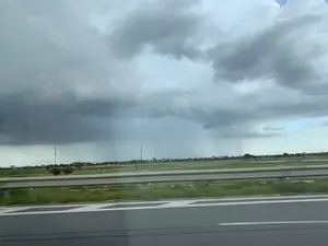 Bức ảnh chụp khung cảnh nhìn từ một phương tiện đang di chuyển trên đường cao tốc. Ở tiền cảnh là mặt đường nhựa, vạch kẻ đường và lan can bảo vệ. Phía bên kia đường là một vùng nông thôn rộng lớn với những cánh đồng xanh. Ở phía xa có thể thấy những ngôi nhà, cây cối và các cột điện. Bầu trời u ám, bị bao phủ bởi những đám mây giông xám xịt, dày đặc, báo hiệu một cơn mưa sắp đến. Có thể quan sát thấy những vệt mưa đang rơi ở phía xa.。（日本語訳）。高速道路を走行中の乗り物から見た風景を撮影した写真です。手前にはアスファルトの路面、車線、ガードレールがあります。道路の向こう側には、緑の田んぼが広がる広大な田園地帯があります。遠くには家々、木々、そして電柱が見えます。空はどんよりとしており、濃く暗い灰色の雷雲に覆われ、雨が間もなく降ることを知らせています。遠くで雨が降っている筋が見えます。