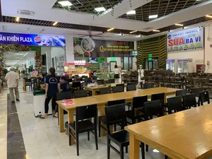 Bối cảnh là một khu vực ăn uống rộng rãi, giống như một khu ẩm thực (food court) bên trong một trạm dừng nghỉ. Không gian được bố trí nhiều bộ bàn ghế gỗ màu nâu và ghế màu đen để phục vụ khách hàng. Có nhiều người trong ảnh, bao gồm cả nhân viên và khách hàng.。Các dòng chữ ghi nhận được từ các biển hiệu và đồng phục:。1.  Biển hiệu lớn bên trái: "XUÂN KHIÊM PLAZA" và logo "PEPSI".。2.  Biển hiệu nhà hàng ở giữa: "RED MOON", "MÓN NGON VÙNG MIỀN", "QUẤY ĂN TỰ CHỌN ĐẬM VỊ QUÊ HƯƠNG". Bên dưới có ghi địa chỉ: "CƠ SỞ 1: NHÀ 16 NGÕ 138 ĐƯỜNG NÚI...", "CƠ SỞ 2: TRẠM DỪNG NGHỈ XUÂN KHIÊM".。3.  Biển quảng cáo lớn bên phải: "CÔNG TY CP SẢN XUẤT SỮA BA VÌ", "OCOP", "SỮA BA VÌ".。4.  Tại quầy bán đồ ăn: "TRẠM DỪNG NGHỈ XUÂN KHIÊM", "BÁNH MÌ 89M", "CHẢ CÁ NHÁY", "NEM CHUA THANH HÓA KHANH LẠN".。5.  Trên lưng áo đồng phục của nhân viên: "XUÂN KHIÊM GROUP".。6.  Biển chỉ dẫn: "WC" (nhà vệ sinh).。Hoạt động đang diễn ra là hoạt động kinh doanh ăn uống tại một trạm dừng nghỉ. Một nhóm nhân viên mặc đồng phục đang đứng gần khu vực phục vụ. Một vài khách hàng đang đi lại trong không gian của khu ẩm thực. Có các quầy hàng đang hoạt động, bán các đặc sản địa phương như nem chua Thanh Hóa và các món ăn khác.。（日本語訳）。背景は、サービスエリア内にあるフードコートのような、広々とした飲食エリアです。空間には、お客様をもてなすため、茶色の木製テーブルと椅子、そして黒い椅子が多数配置されています。看板や制服から読み取れる文字：。1.  左側の大きな看板：「XUÂN KHIÊM PLAZA」と「PEPSI」のロゴ。2.  中央のレストランの看板：「RED MOON」、「MÓN NGON VÙNG MIỀN」（郷土の美味しい料理）、「QUẤY ĂN TỰ CHỌN ĐẬM VỊ QUÊ HƯƠNG」（故郷の味豊かなセルフサービスカウンター）。その下には住所が記載されています：「CƠ SỞ 1: NHÀ 16 NGÕ 138 ĐƯỜNG NÚI...」（1号店：NUI通り138路地16番...）、「CƠ SỞ 2: TRẠM DỪNG NGHỈ XUÂN KHIÊM」（2号店：XUAN KHIEMサービスエリア）。3.  右側の大きな広告看板：「CÔNG TY CP SẢN XUẤT SỮA BA VÌ」（BA VI牛乳生産株式会社）、「OCOP」、「SỮA BA VÌ」（BA VI牛乳）。4.  食べ物販売カウンターにて：「TRẠM DỪNG NGHỈ XUÂN KHIÊM」（XUAN KHIEMサービスエリア）、「BÁNH MÌ 89M」（89Mバインミー）、「CHẢ CÁ NHÁY」（NHAY魚のさつま揚げ）、「NEM CHUA THANH HÓA KHANH LẠN」（KHANH LANのタインホア風ネムチュア）。5.  従業員の制服の背中：「XUÂN KHIÊM GROUP」。6.  案内表示：「WC」（トイレ）。行われている活動は、サービスエリアでの飲食業の営業です。制服を着用した従業員の一団が、提供エリアの近くに立っています。数人のお客様がフードコートの空間内を歩き回っています。タインホアのネムチュアのような地元の特産品やその他の料理を販売している、営業中の売店があります。