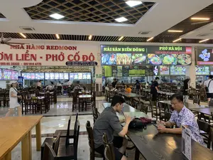 Đây là khung cảnh bên trong một khu vực ẩm thực rộng lớn, có thể là một trạm dừng nghỉ. Không gian có nhiều bàn ghế gỗ dành cho thực khách và nhiều quầy hàng ăn uống với các biển hiệu lớn, sáng đèn.。Các dòng chữ trên biển hiệu có thể đọc được bao gồm:。- "NHÀ HÀNG REDMOON" và "CƠM LIỀN - PHỞ CỐ ĐÔ", kèm số điện thoại "085 65 33333 - 097 919 4775". Địa chỉ được liệt kê: "CƠ SỞ 1 : NHÀ 16 NGÕ 138 ĐƯỜNG NÚI VÀNG - TP TAM ĐIỆP" và "CƠ SỞ 2 : XUÂN KHIÊM PLAZA - CAO TỐC MAI SƠN - CAO BỒ - NB".。- Quầy "HẢI SẢN BIỂN ĐÔNG" với thực đơn các món hải sản.。- Quầy "PHỞ HÀ NỘI" với dòng chữ "THE WORLD'S 50 BEST FOODS" và logo CNN, cùng biển chỉ dẫn "LỐI VÀO LỐI RA".。Hoạt động đang diễn ra là mọi người đang ăn uống và nghỉ ngơi. Ở phía trước, hai người đàn ông đang ngồi tại bàn, uống nước và xem điện thoại. Các khách hàng khác đang ngồi tại các bàn ăn, trong khi một số người đang đi lại trong khu vực. Nhân viên nhà hàng đang làm việc phía sau các quầy hàng. Phía trên một quầy hàng có treo những chiếc nón lá để trang trí.。（日本語訳）。これは広大なフードエリアの内部の光景で、サービスエリアかもしれません。空間には食事客用の木製のテーブルと椅子が多数あり、大きな看板に明かりが灯った多くの飲食店があります。看板に書かれている読み取れる文字には、以下が含まれます：。- 「NHÀ HÀNG REDMOON（レッドムーンレストラン）」と「CƠM LIỀN - PHỞ CỐ ĐÔ（定食 - 古都フォー）」、電話番号「085 65 33333 - 097 919 4775」付き。記載されている住所：「CƠ SỞ 1 : NHÀ 16 NGÕ 138 ĐƯỜNG NÚI VÀNG - TP TAM ĐIỆP（1号店：タムディエップ市、ヌイヴァン通り、138路地、16番）」と「CƠ SỞ 2 : XUÂN KHIÊM PLAZA - CAO TỐC MAI SƠN - CAO BỒ - NB（2号店：スアンキエムプラザ - マイソン-カオボー高速道路 - NB）」。- 海鮮料理のメニューがある「HẢI SẢN BIỂN ĐÔNG（東海シーフード）」のカウンター。- 「THE WORLD'S 50 BEST FOODS」の文字とCNNのロゴ、そして「LỐI VÀO LỐI RA（出入口）」の案内板がある「PHỞ HÀ NỘI（ハノイフォー）」のカウンター。行われている活動は、人々が食事をしたり休憩したりしていることです。前方では、二人の男性がテーブルに座り、飲み物を飲みながら携帯電話を見ています。他の客たちは食事用のテーブルに座っており、一方で何人かの人々はエリア内を歩き回っています。レストランの従業員はカウンターの後ろで働いています。あるカウンターの上には、装飾のためにノンラーがいくつか吊るされています。