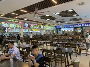 Bối cảnh là một khu ẩm thực (food court) rộng rãi bên trong một tòa nhà, có khả năng là một trạm dừng nghỉ hoặc trung tâm thương mại tên là "XUÂN KHIÊM PLAZA". Không gian được bố trí với rất nhiều dãy bàn ghế gỗ màu sẫm cho thực khách. Trần nhà cao, được trang bị hệ thống đèn chiếu sáng và quạt trần công nghiệp cỡ lớn. Phía sau là các quầy hàng ăn uống liền kề nhau, mỗi quầy đều có biển hiệu lớn được chiếu sáng. Một số người đang ngồi ăn, một số khác đang đi lại hoặc đứng trước các quầy để gọi món. Nhân viên cũng có mặt tại các quầy hàng.。Các dòng chữ có thể đọc được trên các biển hiệu bao gồm:。- Một biển hiệu lớn màu xanh dương ghi "XUÂN KHIÊM PLAZA".。- Một quầy hàng có tên "PHỞ HÀ NỘI" và dòng chữ tiếng Anh "THE WORLD'S 50 BEST FOODS" cùng logo của CNN.。- Một quầy hàng khác tên "NHÀ HÀNG ẨM THỰC 3 MIỀN Hương vị Việt", quảng cáo các món "CƠM TẤM SƯỜN - BÚN BÒ HUẾ - CƠM QUÊ" và có số điện thoại "HOTLINE: 0976232622".。- Một quầy hàng ghi "CƠM QUÊ - DÊ NÚI".。- Một quầy hàng có tên "Nhà hàng Vẹ Vàng" và "Ẩm Thực Bình Pháp".。- Một cửa hàng "Kem Ý tự chọn".。Ở tiền cảnh, hai người đàn ông đang ngồi tại một chiếc bàn. Một người lớn tuổi hơn đang cầm ly nước và nhìn vào điện thoại. Người còn lại trẻ hơn, đang đeo tai nghe và cũng đang sử dụng điện thoại.。（日本語訳）。背景は、ある建物の中にある広々としたフードコートで、「XUÂN KHIÊM PLAZA」という名前のサービスエリアかショッピングセンターの可能性があります。スペースには、食事客のために暗い色の木製のテーブルと椅子が多数並べられています。天井は高く、照明システムと大型の業務用シーリングファンが備え付けられています。後方には飲食の屋台が隣接しており、各屋台には照明付きの大きな看板があります。食事をしている人もいれば、歩き回ったり、注文するために屋台の前に立ったりしている人もいます。屋台にはスタッフもいます。看板で読み取れる文字には、以下が含まれます：。- 「XUÂN KHIÊM PLAZA」と書かれた青い大きな看板。- 「PHỞ HÀ NỘI」という名前の屋台と、英語の「THE WORLD'S 50 BEST FOODS」という文字、そしてCNNのロゴ。- 「NHÀ HÀNG ẨM THỰC 3 MIỀN Hương vị Việt」という名前の別の屋台。「CƠM TẤM SƯỜN - BÚN BÒ HUẾ - CƠM QUÊ」の各料理を宣伝しており、「HOTLINE: 0976232622」という電話番号が記載されている。- 「CƠM QUÊ - DÊ NÚI」と書かれた屋台。- 「Nhà hàng Vẹ Vàng」と「Ẩm Thực Bình Pháp」という名前の屋台。- 「Kem Ý tự chọn」（セルフサービスのイタリアンジェラート）の店。手前では、二人の男性がテーブルに座っています。年配の男性はコップを持ち、スマートフォンを見ています。もう一人の若い男性は、イヤホンをつけ、彼もまたスマートフォンを使っています。