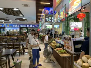 Bức ảnh chụp khung cảnh bên trong một khu ẩm thực hoặc trạm dừng nghỉ hiện đại có tên là "XUÂN KHIÊM PLAZA". Không gian có nhiều bàn ghế gỗ cho khách ngồi và một dãy các quầy hàng ăn uống. Nhiều người, bao gồm cả khách hàng và nhân viên, đang có mặt. Một số khách hàng đang đi lại và lựa chọn đồ ăn, trong khi nhân viên đang đứng sau quầy để phục vụ. Trên các quầy hàng có trưng bày nhiều loại trái cây tươi như dừa, dưa hấu, ổi, táo và các món ăn nhanh như bánh mì.。Các dòng chữ và biển hiệu có thể đọc được bao gồm:。- Tên địa điểm chính: "XUÂN KHIÊM PLAZA".。- Các quầy hàng và sản phẩm: "Kem Ý tự chọn", "Wow。Chè" (với các món Chè Thái, Chè Sâm), "COFFEE - TRÀ SỮA", "NƯỚC ÉP - TRÀ HOA QUẢ", "GÀ RÁN", "Sữa Hạt Mix Vị".。- Một quầy hàng tên "CAFE SEN" bán các loại cà phê (`Café muối`, `Cafe sữa`, `Bạc xỉu`), `NƯỚC GIẢI KHÁT` và `ĐỒ ĂN NHANH`.。- Bảng giá các món ăn: `PATE CHẢ 35.000`, `XÚC XÍCH 25.000`, `THỊT XIÊN 30.000`, `BÁNH BAO 25.000`.。- Biển chỉ dẫn: "KHU VỰC QUẦY THANH TOÁN".。- Quảng cáo: "UỐNG LÀ PHÊ BOSS CÀ PHÊ SỮA".。Các vật dụng khác có thể thấy trong ảnh là quạt điện, lò vi sóng hiệu "BlueStone" và những chiếc đèn lồng đỏ được treo để trang trí.。（日本語訳）。「XUÂN KHIÊM PLAZA」という名前の近代的なフードコートまたは休憩所の内部の光景を撮影した写真です。スペースには、客が座るための木製のテーブルと椅子が多数あり、飲食店が並んでいます。客やスタッフを含む多くの人がいます。一部の客は歩き回って食べ物を選んでおり、一方、スタッフはカウンターの後ろに立って接客しています。各屋台には、ココナッツ、スイカ、グアバ、リンゴなどの新鮮な果物や、バインミーなどの軽食が陳列されています。読み取れる文字や看板は以下の通りです：。- 主な場所の名前：「XUÂN KHIÊM PLAZA」。- 屋台と商品：「セルフサービスイタリアンジェラート」、「Wow。チェー」（タイ風チェー、高麗人参チェーなど）、「COFFEE - TRÀ SỮA（コーヒー - ミルクティー）」、「NƯỚC ÉP - TRÀ HOA QUẢ（ジュース - フルーツティー）」、「GÀ RÁN（フライドチキン）」、「Sữa Hạt Mix Vị（ミックスナッツミルク）」。- 「CAFE SEN」という名前の屋台では、各種コーヒー（`Café muối`（塩コーヒー）、`Cafe sữa`（ミルクコーヒー）、`Bạc xỉu`（バクシウ））、`NƯỚC GIẢI KHÁT`（清涼飲料水）、`ĐỒ ĂN NHANH`（軽食）を販売しています。- 料理の価格表：`PATE CHẢ（パテ・チャー） 35.000`、`XÚC XÍCH（ソーセージ） 25.000`、`THỊT XIÊN（串焼き肉） 30.000`、`BÁNH BAO（肉まん） 25.000`。- 案内表示：「KHU VỰC QUẦY THANH TOÁN（会計カウンターエリア）」。- 広告：「UỐNG LÀ PHÊ BOSS CÀ PHÊ SỮA（ボスミルクコーヒーを飲めば最高）」。写真に見られるその他の物としては、扇風機、「BlueStone」ブランドの電子レンジ、そして装飾のために吊るされた赤い提灯があります。