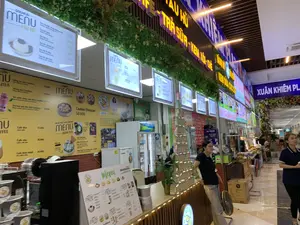 Bối cảnh là một khu ẩm thực trong nhà, có thể là bên trong trung tâm thương mại Xuân Khiêm Plaza, như được chỉ ra bởi một biển hiệu lớn màu xanh ở phía xa. Một dãy các quầy hàng đồ ăn và thức uống được sắp xếp liền kề nhau.。Một quầy hàng nổi bật với biển hiệu lớn màu vàng ghi các món: '...TÀU HŨ - TRÀ SỮA - KEM XÔI - CHÈ'. Phía trên quầy có nhiều bảng menu điện tử sáng đèn. Các văn bản có thể đọc được từ các menu bao gồm:。- Menu 'DOUHUA - TÀU HŨ' liệt kê các món: Tàu hũ trân châu đường đen (35.000), Tàu hũ xoài (35.000), Tàu hũ bơ (35.000), Tàu hũ chanh leo (35.000), Tàu hũ lá nếp (35.000), Tàu hũ dâu tằm (35.000).。- Menu đồ uống và đồ ăn khác: Hồng trà sữa ComAtea (50.000), Olong sữa ComAtea (50.000), Cafe đen (25.000), Cafe nâu (30.000), Combo Dimsum (50.000), và menu 'KEM XÔI' với các món như Chè Cốm Bưởi (40.000) và Kem Xôi Dừa Socola/Bạc Hà Trà Xanh (25.000).。Hoạt động trong ảnh là cảnh kinh doanh thường thấy tại một khu ăn uống, với một nhân viên nữ đang đứng tại quầy và một vài khách hàng đang đi lại. Các vật dụng có thể thấy bao gồm tủ lạnh, một tháp lon nước được xếp để trưng bày, quạt điện đặt trên sàn, và các hộp carton.。（日本語訳）。背景は屋内のフードコートで、遠くにある大きな青い看板が示すように、おそらくスアンキエムプラザのショッピングセンター内です。食べ物や飲み物の屋台が隣り合って並んでいます。ある屋台は、黄色い大きな看板が目立ち、メニューが書かれています：「...豆腐花 - ミルクティー - アイスクリームともち米 - チェー」。カウンターの上には、ライトアップされた電子メニューボードが多数あります。メニューから読み取れるテキストには以下が含まれます：。- 「DOUHUA - 豆腐花」メニューには以下の品目がリストされています：黒糖タピオカ豆腐花（35,000）、マンゴー豆腐花（35,000）、アボカド豆腐花（35,000）、パッションフルーツ豆腐花（35,000）、パンダンリーフ豆腐花（35,000）、マルベリー豆腐花（35,000）。- その他の飲み物と食べ物のメニュー：ComAtea紅茶ミルクティー（50,000）、ComAteaウーロンミルクティー（50,000）、ブラックコーヒー（25,000）、ブラウンコーヒー（30,000）、点心コンボ（50,000）、そして「アイスクリームともち米」メニューには、緑米とザボンのチェー（40,000）やココナッツもち米アイスクリーム チョコレート/ミント緑茶（25,000）のような品目があります。写真の中の活動は、フードコートでよく見られる営業風景で、女性スタッフがカウンターに立っており、数人の客が歩き回っています。