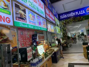 Bức ảnh chụp tại khu vực ẩm thực bên trong một trung tâm thương mại hoặc quảng trường, có thể là 'XUÂN KHIÊM PLAZA' như trên biển hiệu. Không gian là một hành lang rộng rãi với sàn gạch bóng, có nhiều quầy hàng đồ ăn và thức uống được chiếu sáng.。Các thông tin bằng chữ có thể nhận dạng bao gồm:。- Biển hiệu lớn: 'XUÂN KHIÊM PLAZA' và logo Pepsi.。- Biển hiệu các quầy hàng và sản phẩm: 'SỮA CHUA TRÂN CHÂU HẠ LONG', 'KEM - SỮA CHUA', 'KEM TRÁI CÂY CHÀO HÈ。', 'Cà Phê Muối', 'Thanh Nhiệt - Giải Độc - Giảm Cân', 'Doner kebab', 'CAY - TOKBOKKI', 'ĐỒ ĂN NHANH', 'CÀ PHÊ MÁY'.。- Biển chỉ dẫn: 'WC' (nhà vệ sinh).。- Thực đơn (Menu) liệt kê các món như Cà Phê Đen, Trà Chanh, Nước Ép Hoa Quả, Sữa Chua, Ca Cao với giá tiền đi kèm.。Hoạt động chính trong ảnh là một người phụ nữ đang đứng tại một quầy hàng, có thể đang lựa chọn hoặc chờ mua đồ uống. Các vật thể nổi bật bao gồm nhiều loại trái cây tươi như dừa, dứa, chanh được bày bán; một máy pha cà phê; một tủ đông kem hiệu 'Aice'; các chai nước khoáng và lon nước ngọt; đồ ăn nhanh trong tủ kính. Lối đi được trang trí bằng một cổng hoa lớn nhiều màu sắc.。（日本語訳）。写真は、ショッピングセンターまたは広場内のフードコートエリアで撮影されたもので、看板にあるように「XUÂN KHIÊM PLAZA」である可能性があります。空間は、光沢のあるタイル張りの床を持つ広々とした通路で、照明で照らされた多くの飲食物の屋台があります。識別できる文字情報には以下が含まれます：。- 大きな看板：「XUÂN KHIÊM PLAZA」とペプシのロゴ。- 各屋台と商品の看板：「SỮA CHUA TRÂN CHÂU HẠ LONG」（ハロン・タピオカヨーグルト）、「KEM - SỮA CHUA」（アイスクリーム - ヨーグルト）、「KEM TRÁI CÂY CHÀO HÈ。」（夏を迎えるフルーツアイス。）、「Cà Phê Muối」（塩コーヒー）、「Thanh Nhiệt - Giải Độc - Giảm Cân」（熱冷まし - デトックス - ダイエット）、「Doner kebab」（ドネルケバブ）、「CAY - TOKBOKKI」（辛い - トッポギ）、「ĐỒ ĂN NHANH」（ファストフード）、「CÀ PHÊ MÁY」（マシンコーヒー）。- 案内標識：「WC」（トイレ）。- メニュー：ブラックコーヒー、レモンティー、フルーツジュース、ヨーグルト、ココアなどの品目が価格と共に記載されています。写真の中の主な活動は、一人の女性が屋台に立っており、飲み物を選んでいるか、購入を待っている可能性があります。目立つ物には、販売用に陳列されているココナッツ、パイナップル、レモンなどの多くの種類の新鮮な果物、コーヒーマシン1台、「Aice」ブランドのアイスクリーム冷凍庫1台、ミネラルウォーターのボトルとソフトドリンクの缶、ガラスケースの中のファストフードが含まれます。通路は、大きな色とりどりの花のアーチで飾られています。