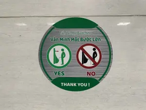Đây là hình ảnh một nhãn dán hình tròn được dán trên tường hoặc sàn gạch men, có khả năng là trong một nhà vệ sinh nam. Trên nhãn dán có các dòng chữ tiếng Việt và tiếng Anh. Dòng chữ tiếng Việt là "Tiến Lên Một Bước Nhỏ, Văn Minh Một Bước Lớn". Bên dưới là hai biểu tượng: một biểu tượng trong vòng tròn màu xanh lá cây với chữ "YES", mô tả hành động đúng là một người đứng gần bồn tiểu khi đi vệ sinh; và một biểu tượng cấm trong vòng tròn màu đỏ với chữ "NO", mô tả hành động sai là đứng xa bồn tiểu. Dưới cùng là dòng chữ "THANK YOU。". Mục đích của nhãn dán này là để hướng dẫn và nhắc nhở mọi người giữ gìn vệ sinh chung tại khu vực toilet.。（日本語訳）。これは、壁またはタイル張りの床に貼られた円形のステッカーの画像で、おそらく男子トイレの中のものでしょう。ステッカーにはベトナム語と英語の文言があります。ベトナム語の文言は「Tiến Lên Một Bước Nhỏ, Văn Minh Một Bước Lớn」です。その下には2つのアイコンがあります。1つは「YES」という文字が入った緑色の円の中のアイコンで、用を足す際に小便器の近くに立つという正しい行動を描写しています。そして、もう1つは「NO」という文字が入った赤色の円の中の禁止アイコンで、小便器から離れて立つという誤った行動を描写しています。一番下には「THANK YOU。」という文言があります。このステッカーの目的は、トイレエリアの公衆衛生を保つよう、人々に案内し注意を促すことです。