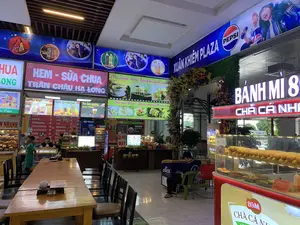 Đây là khu vực ăn uống trong nhà tại một trung tâm thương mại hoặc trạm dừng nghỉ có tên là Xuân Khiêm Plaza. Không gian được chiếu sáng rực rỡ bởi nhiều biển hiệu đèn của các quầy hàng.。Các dòng chữ có thể đọc được bao gồm:。- Tên khu vực: "XUÂN KHIÊM PLAZA".。- Các quầy hàng: "BÁNH MÌ 89 CHẢ CÁ NH...", "KEM - SỮA CHUA TRÂN CHÂU HẠ LONG", quầy bán "Cà Phê - Trà sữa - Sinh tố - Nước ép trái cây tươi nguyên" và "Thế giới đồ ăn – Thiên đường đồ uống".。- Menu đồ uống: "Cà Phê Muối", "Coffee Bạc Sỉu", "Trà Sữa Nướng kem trứng", "Nước Ép Hoa Quả", "Trà Sữa Trân Châu Đường Đen", "TOPPING TRÂN CHÂU TRẮNG, KEM CHEESE".。- Biển quảng cáo: "PEPSI", "STING BẬT NĂNG LƯỢNG SỐNG BỨT PHÁ", "BOSS COFFEE UỐNG LÀ PHÊ", "TEA+".。Hoạt động đang diễn ra là kinh doanh đồ ăn và thức uống. Có một vài khách hàng trong khu vực: một người đàn ông đang ngồi xem điện thoại tại quầy, một người khác ngồi trên ghế nhựa gần tủ đông "Aice", và một cậu bé đang đi lại. Các quầy hàng trưng bày sản phẩm như bánh mì trong tủ kính, dừa tươi và các loại đồ uống khác. Có nhiều bàn ghế gỗ dài được sắp xếp cho khách ngồi.。（日本語訳）。これは、スアンキエムプラザという名前のショッピングセンターまたはサービスエリアにある屋内の飲食エリアです。空間は、各屋台の多くの電飾看板によって明るく照らされています。読み取れる文字は以下の通りです：。- エリア名：「XUÂN KHIÊM PLAZA」。- 屋台：「BÁNH MÌ 89 CHẢ CÁ NH...」、「KEM - SỮA CHUA TRÂN CHÂU HẠ LONG」、そして「Cà Phê - Trà sữa - Sinh tố - Nước ép trái cây tươi nguyên」（コーヒー - ミルクティー - スムージー - 生搾りフルーツジュース）と「Thế giới đồ ăn – Thiên đường đồ uống」（食べ物の世界 – 飲み物の天国）を販売する屋台。- ドリンクメニュー：「Cà Phê Muối」（塩コーヒー）、「Coffee Bạc Sỉu」（バクシウコーヒー）、「Trà Sữa Nướng kem trứng」（焼きミルクティーエッグクリーム）、「Nước Ép Hoa Quả」（フルーツジュース）、「Trà Sữa Trân Châu Đường Đen」（黒糖タピオカミルクティー）、「TOPPING TRÂN CHÂU TRẮNG, KEM CHEESE」（トッピング 白タピオカ、クリームチーズ）。- 広告看板：「PEPSI」、「STING BẬT NĂNG LƯỢNG SỐNG BỨT PHÁ」、「BOSS COFFEE UỐNG LÀ PHÊ」、「TEA+」。行われている活動は、飲食物の販売です。エリア内には数人の客がいます：カウンターで携帯電話を見ている男性が一人、冷凍庫「Aice」の近くのプラスチック椅子に座っている別の人が一人、そして歩き回っている男の子が一人います。各屋台では、ガラスケースの中のバインミー、新鮮なココナッツ、その他の各種飲み物などの商品が陳列されています。客が座れるように、多くの長い木製のテーブルと椅子が配置されています。