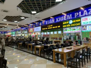 Bức ảnh chụp khung cảnh bên trong một khu ẩm thực (food court) rộng rãi, có tên là 'XUÂN KHIÊM PLAZA' được ghi trên biển hiệu lớn màu xanh dương. Đây là một không gian trong nhà với sàn lát gạch men sáng bóng và nhiều bộ bàn ghế gỗ được sắp xếp ngay ngắn cho thực khách. Phía trên trần nhà có trang trí giàn cây leo nhân tạo.。Dọc theo khu vực này là một dãy các quầy hàng đồ ăn và thức uống liền kề nhau. Mỗi quầy đều có biển hiệu đèn LED rực rỡ với các món ăn được quảng cáo. Một vài người, bao gồm nhân viên và khách hàng, đang có mặt tại các quầy. Hầu hết các bàn đều trống.。Các dòng chữ trên các biển hiệu có thể nhận dạng được bao gồm:。- Tên khu vực: XUÂN KHIÊM PLAZA.。- Tên các món ăn và đồ uống tại các quầy:。- 'Wow。Chè Ngon'。- '桑的店' (một cửa hàng có tên tiếng Trung), 'TRÀ SỮA, CÀ PHÊ, NƯỚC HOA QUẢ'。- 'V TÀU HŨ', 'TÀU HŨ - CAFÉ - TRÀ SỮA - KEM XÔI - CHÈ'。- 'BÁNH MỲ', 'CHÈ NGON', 'KEM DỪA CUCCUC'。- 'KEM - SỮA CHUA HA LONG' với các món 'CÀ PHÊ', 'SỮA CHUA', 'KEM XÔI', 'KEM CHIÊN'。- 'KEM - TRÂN CHÂU', 'Cà Phê Muối', 'Thanh Nhiệt', 'CÀ PHÊ MÁY'。- Một tấm biển nhỏ đặt trên bàn ghi: 'TRÀ ĐÁ, TRÀ NÓNG, NHÂN TRẦN'.。（日本語訳）。写真は、「XUÂN KHIÊM PLAZA」という名前が大きな青い看板に書かれた、広々としたフードコートの内部の光景を写しています。これは屋内の空間で、床は光沢のあるタイル張りで、食事客のために多くの木製のテーブルと椅子のセットが整然と並べられています。天井には人工のつる植物の棚が飾られています。このエリアに沿って、食べ物と飲み物の屋台が隣接して並んでいます。各屋台には、宣伝されている料理が書かれた鮮やかなLED看板があります。スタッフや客を含む何人かの人々が、各屋台にいます。ほとんどのテーブルは空いています。識別可能な看板の文字には、以下のものが含まれます：。- エリア名：XUÂN KHIÊM PLAZA。- 各屋台の食べ物と飲み物の名前：。- 「Wow。Chè Ngon」（美味しいチェー）。- 「桑的店」（中国語名の店）、「TRÀ SỮA, CÀ PHÊ, NƯỚC HOA QUẢ」（ミルクティー、コーヒー、フルーツジュース）。- 「V TÀU HŨ」、「TÀU HŨ - CAFÉ - TRÀ SỮA - KEM XÔI - CHÈ」（豆花 - カフェ - ミルクティー - もち米アイス - チェー）。- 「BÁNH MỲ」（バインミー）、「CHÈ NGON」（美味しいチェー）、「KEM DỪA CUCCUC」（CUCCUCココナッツアイス）。- 「KEM - SỮA CHUA HA LONG」（ハロン・アイス - ヨーグルト）、メニューは「CÀ PHÊ」（コーヒー）、「SỮA CHUA」（ヨーグルト）、「KEM XÔI」（もち米アイス）、「KEM CHIÊN」（揚げアイス）。- 「KEM - TRÂN CHÂU」（アイス - タピオカ）、「Cà Phê Muối」（塩コーヒー）、「Thanh Nhiệt」（清熱茶）、「CÀ PHÊ MÁY」（マシンコーヒー）。- テーブルの上に置かれた小さな看板には、「TRÀ ĐÁ, TRÀ NÓNG, NHÂN TRẦN」（アイスティー、ホットティー、ニガハッカ茶）と書かれています。