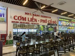 Chữ viết được ghi lại:。- Biển hiệu chính lớn: NHÀ HÀNG REDMOON, CƠM LIỀN - PHỞ CỐ ĐÔ. HOTLINE: 085 65 33333 - 097 919 4775.。- Biển hiệu địa chỉ: CƠ SỞ 1: NHÀ 16 NGÕ 138 ĐƯỜNG NÚI VÀNG TAM ĐIỆP. CƠ SỞ 2: XUÂN KHIÊM PLAZA - CAO TỐC MAI SƠN...。- Biển hiệu các quầy hàng: HẢI SẢN BIỂN ĐÔNG, PHỞ HÀ NỘI, Nhà Hàng Dê Vàng, Ẩm Thực Bình Pháp, THE WORLD'S 50 BEST REST... (chữ bị cắt).。- Các biển hiệu và thông tin khác: BA VÌ, HỘP CHỮA CHÁY, WC, CÓ HÓA ĐƠN VAT, HẾT LÒNG SẼ HẾT TIỀN, Pepsi.。Mô tả bối cảnh và hoạt động:。- Loại địa điểm: Đây là một khu vực ẩm thực trong nhà rộng lớn, giống như một khu ăn uống (food court) hoặc một nhà hàng lớn bên trong một trạm dừng nghỉ trên cao tốc, được xác định qua biển hiệu "XUÂN KHIÊM PLAZA - CAO TỐC MAI SƠN".。- Hoạt động cụ thể: Nhiều thực khách đang ngồi tại các bàn ăn. Nhân viên nhà hàng, một số mặc đồng phục áo phông màu xanh, đang đi lại phục vụ và làm việc tại các quầy chế biến thức ăn. Một người phụ nữ đang ngồi nghỉ ở một chiếc bàn gần lối đi.。- Vật thể trong ảnh: Không gian có rất nhiều bàn ghế gỗ màu nâu sẫm. Trên trần nhà có một chiếc quạt trần công nghiệp lớn đang quay, hệ thống đèn chiếu sáng và điều hòa âm trần. Có nhiều quầy hàng ăn với các biển hiệu đèn LED hiển thị thực đơn, ví dụ như quầy "HẢI SẢN BIỂN ĐÔNG" và "PHỞ HÀ NỘI". Một quầy được trang trí bằng nhiều chiếc nón lá treo trên mái lá nhân tạo. Ngoài ra còn có quạt cây, hộp cứu hỏa màu đỏ, và tủ lạnh đựng nước giải khát.。（日本語訳）。書き記された文字：。- 大きなメイン看板：REDMOONレストラン、CƠM LIỀN - PHỞ CỐ ĐÔ。ホットライン：085 65 33333 - 097 919 4775。- 住所の看板：1号店：タムディップ市ヌイヴァン通り138路地16番。2号店：スアンキエムプラザ - マイソン高速道路...。- 各屋台の看板：東海シーフード、ハノイフォー、Dê Vàng（黄金のヤギ）レストラン、Bình Pháp（ビンファップ）料理、THE WORLD'S 50 BEST REST...（文字が途切れている）。- その他の看板と情報：BA VÌ（バヴィ）、消火器ボックス、WC（トイレ）、VATインボイス発行可、HẾT LÒNG SẼ HẾT TIỀN（心を尽くせば金が尽きる）、Pepsi（ペプシ）。背景と活動の説明：。- 場所の種類：これは広大な屋内の飲食エリアで、フードコートや高速道路のパーキングエリア内にある大きなレストランのようであり、「スアンキエムプラザ - マイソン高速道路」という看板によって特定される。- 具体的な活動：多くの客がテーブル席に座っている。レストランの従業員は、一部は青いTシャツの制服を着ており、行き来しながら接客したり、調理カウンターで作業したりしている。一人の女性が通路近くのテーブルで休んでいる。- 写真の中の物体：空間には濃い茶色の木製のテーブルと椅子がたくさんある。天井には大きな業務用のシーリングファンが回っており、照明システムと天井埋め込み式のエアコンがある。メニューを表示するLED看板が付いた多くの飲食店があり、例えば「東海シーフード」や「ハノイフォー」の屋台がある。ある屋台は、人工の葉葺き屋根にたくさんのノンラー（ベトナムの伝統的な笠）を吊るして装飾されている。その他に、扇風機、赤い消火器ボックス、飲料用の冷蔵庫もある。