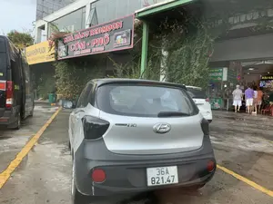 Bối cảnh là một bãi đỗ xe phía trước một khu phức hợp nhà hàng hoặc trạm dừng nghỉ. Nền xi măng ẩm ướt có các vạch sơn màu vàng. Một chiếc ô tô Hyundai Grand i10 màu bạc đang đỗ ở tiền cảnh. Phía sau là tòa nhà có nhiều biển hiệu. Có một biển hiệu màu vàng ghi 'XUÂN KHIÊM PLAZA' và 'KÍNH CHÀO QUÝ KHÁCH。'. Một biển hiệu lớn màu đen-đỏ của 'NHÀ HÀNG REDMOON' quảng cáo 'CƠM - PHỞ' và ghi số điện thoại 'HOTLINE: 085 65 33333 097 919 4775'. Các biển hiệu khác quảng cáo đặc sản như 'DÊ NINH BÌNH TƯƠI NGUYÊN CHẤT' và 'HẢI SẢN'. Dưới mái hiên, một nhóm người đang đứng trước các quầy hàng, có vẻ là khách hàng đang lựa chọn hoặc chờ đợi đồ ăn.。（日本語訳）。背景は、レストラン複合施設またはサービスエリアの前にある駐車場です。湿ったセメントの地面には黄色い塗装線があります。前景には銀色のヒュンダイ・グランドi10が1台駐車しています。後方には、多くの看板がある建物があります。黄色い看板があり、「XUÂN KHIÊM PLAZA」と「KÍNH CHÀO QUÝ KHÁCH。」（お客様、ようこそ。）と書かれています。「NHÀ HÀNG REDMOON」の大きな黒と赤の看板は、「CƠM - PHỞ」（ご飯 - フォー）を宣伝し、電話番号「HOTLINE: 085 65 33333 097 919 4775」が記載されています。他の看板は、「DÊ NINH BÌNH TƯƠI NGUYÊN CHẤT」（新鮮で純粋なニンビン省のヤギ肉）や「HẢI SẢN」（海産物）のような特産品を宣伝しています。軒下では、一団の人々が屋台の前に立っており、食べ物を選んだり待ったりしている客のようです。