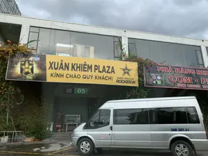 Bức ảnh chụp phía trước một tòa nhà thương mại hiện đại có tên là Xuân Khiêm Plaza. Trên tòa nhà có treo các biển hiệu lớn. Một biển hiệu màu vàng ghi "XUÂN KHIÊM PLAZA" và "KÍNH CHÀO QUÝ KHÁCH。", đồng thời quảng cáo cho "NƯỚC TĂNG LỰC ROCKSTAR" với dòng chữ "THƯƠNG HIỆU ĐẾN TỪ MỸ". Bên cạnh là biển hiệu của một nhà hàng với dòng chữ "NHÀ HÀNG RED...", "CƠM - PH..." và số điện thoại "HOTLINE: 085 6533333 097 9...". Bên dưới có biển chỉ dẫn "CỬA GATE 05". Một chiếc xe ô tô 16 chỗ màu bạc đang đỗ ở phía trước. Khung cảnh là một bãi đỗ xe hoặc lối vào, mặt đất ẩm ướt, tường nhà có giàn hoa giấy và bầu trời u ám.。（日本語訳）。スアンキエムプラザという名前の近代的な商業ビルの前で撮影された写真です。建物には大きな看板が掲げられています。黄色い看板には「XUÂN KHIÊM PLAZA」と「KÍNH CHÀO QUÝ KHÁCH。」と書かれており、同時に「THƯƠNG HIỆU ĐẾN TỪ MỸ」という文言で「NƯỚC TĂNG LỰC ROCKSTAR」を宣伝しています。隣には、「NHÀ HÀNG RED...」、「CƠM - PH...」という文字と、「HOTLINE: 085 6533333 097 9...」という電話番号が書かれたレストランの看板があります。下には「CỬA GATE 05」という案内表示があります。前方に銀色の16人乗りの車が1台停まっています。駐車場または入口の光景で、地面は濡れており、建物の壁にはブーゲンビリアの棚があり、空は曇っています。