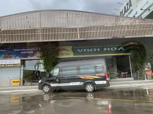 Bức ảnh chụp một khu vực dường như là một trạm dừng nghỉ ven đường, với một bãi đậu xe bị ướt sau cơn mưa, có những vũng nước phản chiếu hình ảnh. Một chiếc xe khách loại nhỏ (minibus) màu đen, có chữ 'DCAR' trên thân, đang đỗ trước một dãy cửa hàng. Bên trong một quán ăn đang mở, có một vài người khách đang ngồi tại bàn. Các biển hiệu có thể đọc được bao gồm: Biển hiệu lớn của cơ sở 'VINH HOA' quảng cáo 'ĐẶC SẢN NEM CHUA THANH HÓA - CƠ SỞ SẢN XUẤT NEM CHUA' với khẩu hiệu 'CẢM NHẬN SỰ ĐẶC BIỆT - ĂN LÀ NHỚ'. Địa chỉ được ghi là 'Cs1: 228 Ngọc Mai - TP. Thanh Hóa - Cs2: Trạm Dừng Nghỉ Xuân Khiêm' và số điện thoại 'Hotline: 0397 217 403 - 0985 063 823'. Một biển hiệu màu xanh dương có chữ 'TRẠM DỪNG NGHỈ ĐỘNG NINH BÌNH' và quảng cáo đồ uống 'NHO DÂU'. Biển hiệu màu vàng của quán '...ÔNG LÊ NHÂN & CAFE' liệt kê các sản phẩm: 'TÀO PHỚ ĐÀI LOAN', 'TRÀ CLONG', 'CAFE ORGANIC', 'SINH TỐ NƯỚC ÉP', 'QUÀ TẶNG ĐẶC SẢN'. Một biển hiệu nhỏ màu vàng trên tường có chữ 'Meko TO GO'. Một biển hiệu dọc quảng cáo 'PHỞ HÀ NỘI', 'PHỞ GÀ' và '...NG VỊ HÀ NỘI XƯA'. Trên nóc tòa nhà, có một biển quảng cáo lớn với chữ 'PLAZA' và thương hiệu nước uống 'AQUAFINA' cùng dòng chữ '...của sự tinh khiết'.。（日本語訳）。写真は、道路脇の休憩所のようである一帯を撮影したもので、雨上がりの濡れた駐車場には、景色を反射する水たまりができています。車体に「DCAR」と書かれた黒い小型バス（ミニバス）が、店舗が並ぶ前に駐車しています。開いている食堂の中には、数人の客がテーブルに座っています。読み取れる看板には以下のものが含まれます：「VINH HOA」という店の大きな看板には、「タインホア名物ネムチュア - ネムチュア製造所」と広告されており、「特別な味を感じて - 食べたら忘れられない」というスローガンが掲げられています。住所は「支店1：タインホア市ゴックマイ228番地 - 支店2：スアンキエム休憩所」、電話番号は「ホットライン：0397 217 403 - 0985 063 823」と記載されています。青い看板には「ドンニンビン休憩所」と書かれ、「NHO DÂU（ブドウイチゴ）」という飲み物を宣伝しています。「...ÔNG LÊ NHÂN & CAFE」という店の黄色い看板には、「台湾豆花」、「烏龍茶」、「オーガニックコーヒー」、「スムージー・ジュース」、「特産品ギフト」といった商品がリストアップされています。壁には「Meko TO GO」と書かれた小さな黄色い看板があります。縦長の看板には、「ハノイフォー」、「鶏肉のフォー」、そして「...昔のハノイの味」と広告されています。建物の屋上には、「PLAZA」という文字と飲料水ブランド「AQUAFINA」の大きな広告看板があり、「...の純粋さ」という文言が添えられています。