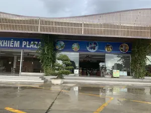 Bức ảnh chụp mặt tiền của một tòa nhà, có vẻ là một trạm dừng nghỉ hoặc trung tâm thương mại nhỏ, có tên là "KHIÊM PLAZA" được ghi trên một biển hiệu lớn màu xanh. Tòa nhà có kiến trúc hiện đại với mặt tiền bằng kính và phần mái hiên được trang trí bằng các thanh gỗ. Phía trước tòa nhà là một khoảng sân bê tông bị ướt, có các vạch kẻ màu vàng, cho thấy trời có thể vừa mưa. Một chậu cây cảnh bonsai được đặt ở phía trước. Nhìn qua cửa kính, có thể thấy bàn ghế bên trong, cho thấy đây là khu vực nghỉ ngơi hoặc ăn uống. Bầu trời trong ảnh u ám và nhiều mây.。（日本語訳）。建物の正面を撮影した写真で、大きな青い看板に「KHIÊM PLAZA」と書かれており、サービスエリアか小さなショッピングセンターのようです。建物は現代的な建築様式で、ガラス張りの正面と木製の梁で装飾された庇があります。建物の前には濡れたコンクリートの広場があり、黄色い線が引かれていて、雨が降ったばかりのようです。前には盆栽の鉢植えが置かれています。ガラスのドア越しに、中のテーブルと椅子が見え、ここが休憩エリアまたは飲食エリアであることを示しています。写真の空はどんよりと曇っています。