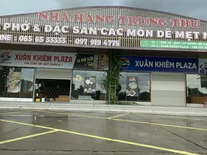 Bức ảnh chụp mặt tiền của một khu phức hợp thương mại, dường như là một trạm dừng nghỉ có tên là Xuân Khiêm Plaza. Phía trên cùng của tòa nhà là một tấm biển lớn với dòng chữ 'NHÀ HÀNG TRUNG THU', quảng cáo các món ăn như 'PHỞ & ĐẶC SẢN CÁC MÓN DÊ MẸT'. Các số điện thoại hotline được liệt kê là '085 65 33333 - 097 919 4775'. Một biển hiệu nhỏ hơn màu xanh lá cây cung cấp thông tin địa chỉ cho hai cơ sở: 'CS 1: NHÀ 16 - NGÕ 138 ĐƯỜNG NÚI VÀ...' và 'CS 2: XUÂN KHIÊM PLAZA - CAO TỐC...'.。Tầng trệt bao gồm nhiều cửa hàng. Các biển hiệu cho thấy đây là 'XUÂN KHIÊM PLAZA' và 'TRẠM DỪNG NGHỈ XUÂN KHIÊM'. Một số cửa hàng có cửa cuốn kim loại đang đóng, trong khi những cửa hàng khác có cửa kính trưng bày các áp phích quảng cáo đồ ăn và thức uống như 'CÀ PHÊ SỮA', 'Bún Chả Quạt' và logo của Pepsi. Một tấm biển ghi 'XIN CẢM ƠN QUÝ KHÁCH。'.。Phía trước tòa nhà là một bãi đỗ xe trải nhựa với các vạch kẻ màu vàng. Mặt đất ướt, cho thấy trời vừa mưa. Khung cảnh vắng lặng, không có hoạt động của người.。（日本語訳）。この写真は、スアンキエム・プラザという名前のサービスエリアと思われる複合商業施設の正面を撮影したものです。建物の最上部には「NHÀ HÀNG TRUNG THU」という文字が書かれた大きな看板があり、「PHỞ & ĐẶC SẢN CÁC MÓN DÊ MẸT」のような料理を宣伝しています。ホットラインの電話番号として「085 65 33333 - 097 919 4775」が記載されています。緑色の小さな看板には、2つの施設の住所情報が記載されています。「CS 1: NHÀ 16 - NGÕ 138 ĐƯỜNG NÚI VÀ...」と「CS 2: XUÂN KHIÊM PLAZA - CAO TỐC...」です。1階には多くの店舗が入っています。看板には「XUÂN KHIÊM PLAZA」と「TRẠM DỪNG NGHỈ XUÂN KHIÊM」と書かれています。いくつかの店舗は金属製のシャッターが閉まっていますが、他の店舗はガラスのドアで、「CÀ PHÊ SỮA」、「Bún Chả Quạt」、ペプシのロゴといった食べ物や飲み物の広告ポスターが飾られています。ある看板には「XIN CẢM ƠN QUÝ KHÁCH。」と書かれています。建物の前には、黄色い線が引かれたアスファルトの駐車場があります。地面は濡れており、雨が降ったばかりであることを示しています。人の気配はなく、静かな光景です。
