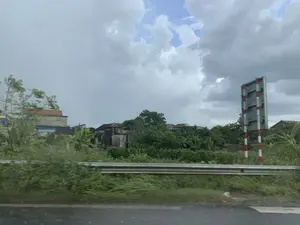 Khung cảnh ven đường ở một vùng nông thôn Việt Nam, được chụp từ một phương tiện đang di chuyển. Con đường nhựa ở phía trước có vẻ ẩm ướt. Dọc bên đường là một hàng rào hộ lan bằng kim loại và cây cỏ mọc um tùm. Ở phía sau, có thể thấy nhiều ngôi nhà dân với mái ngói đặc trưng của làng quê, nằm xen lẫn giữa những lùm cây xanh, bao gồm cả cây chuối. Bên phải hình ảnh có một biển báo giao thông hình chữ nhật, nhưng chỉ nhìn thấy mặt sau của nó. Các cột đỡ biển báo được sơn sọc đỏ và trắng. Bầu trời đầy mây, cho thấy thời tiết có thể sắp mưa hoặc vừa tạnh mưa. Không có hoạt động nào của con người được ghi lại trong ảnh.。（日本語訳）。ベトナムの田舎の道端の風景で、移動中の乗り物から撮影されたものです。前方のアスファルト道路は濡れているように見えます。道端には金属製のガードレールと、生い茂る草木があります。後方には、田舎特有の瓦屋根を持つ民家が、バナナの木を含む緑の木々の間に点在しているのが見えます。画像の右側には長方形の交通標識がありますが、その裏側しか見えません。標識を支える柱は赤と白の縞模様に塗られています。空は雲に覆われており、これから雨が降るか、あるいは雨が上がったばかりであることを示しています。写真には人間の活動は記録されていません。